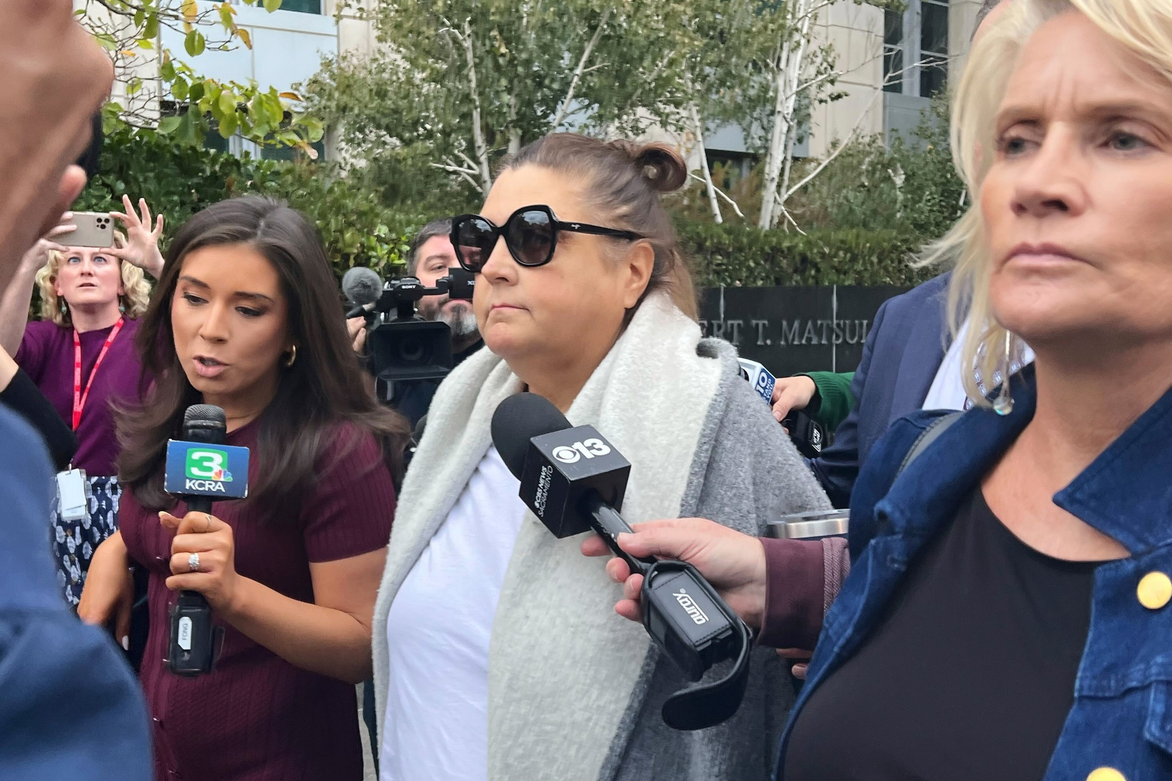 A woman wearing sunglasses and a white scarf is surrounded by reporters holding microphones from news channels KCRA and CBS 13.