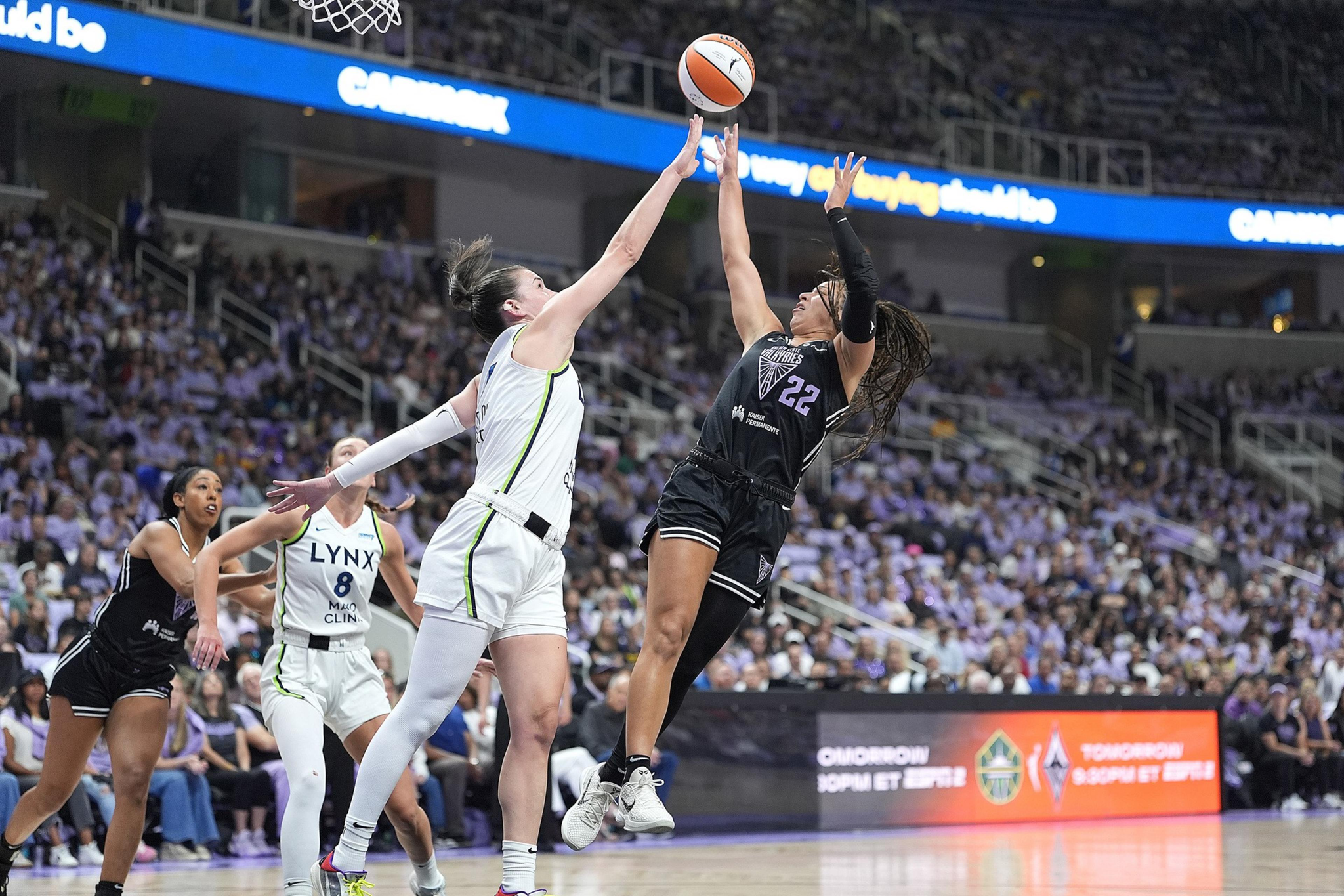 Two women basketball players leap, reaching for the ball mid-air, with one in black and the other in white, while a crowd watches in the background.