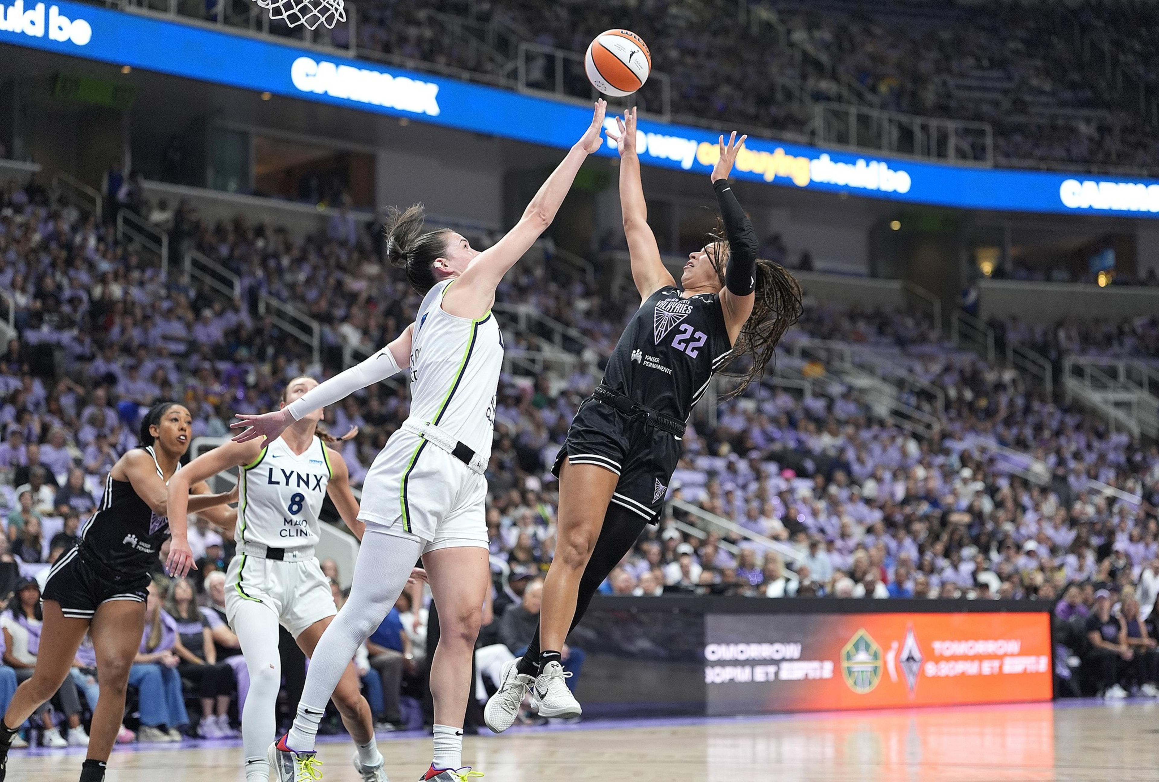 Two women basketball players leap, reaching for the ball mid-air, with one in black and the other in white, while a crowd watches in the background.