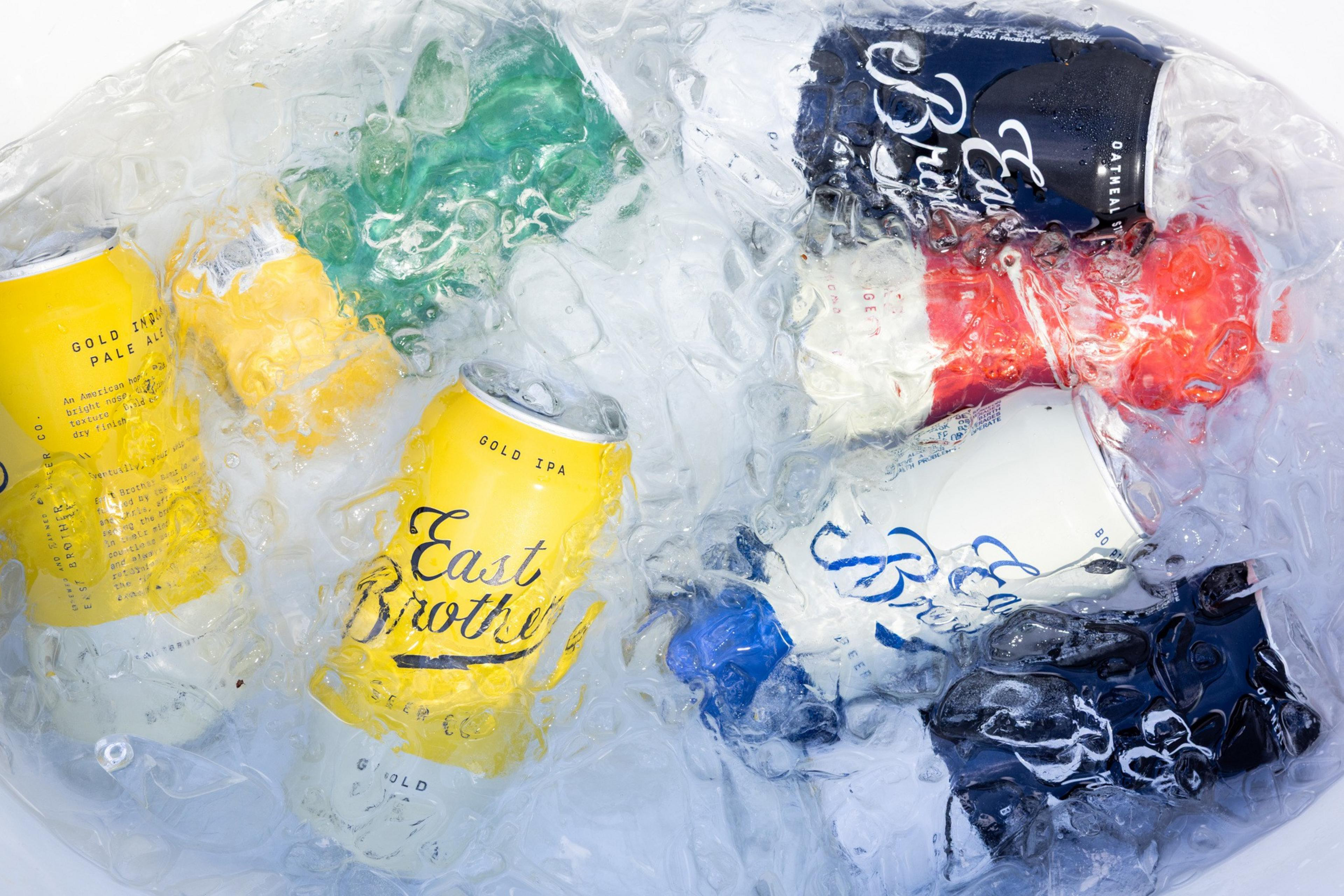 Several colorful beverage cans, including yellow, green, blue, red, and white ones, are submerged in ice inside a cooler or container.
