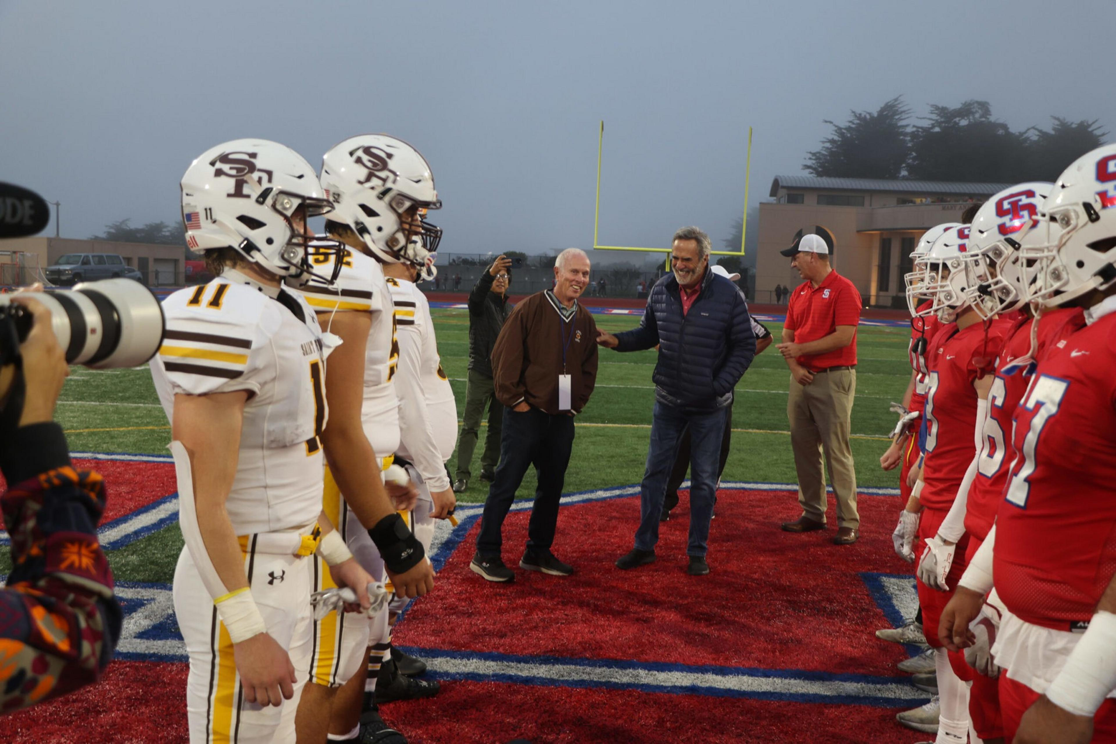 Two football teams in white and red uniforms face each other on a field, with two men standing and talking on the red turf between them.