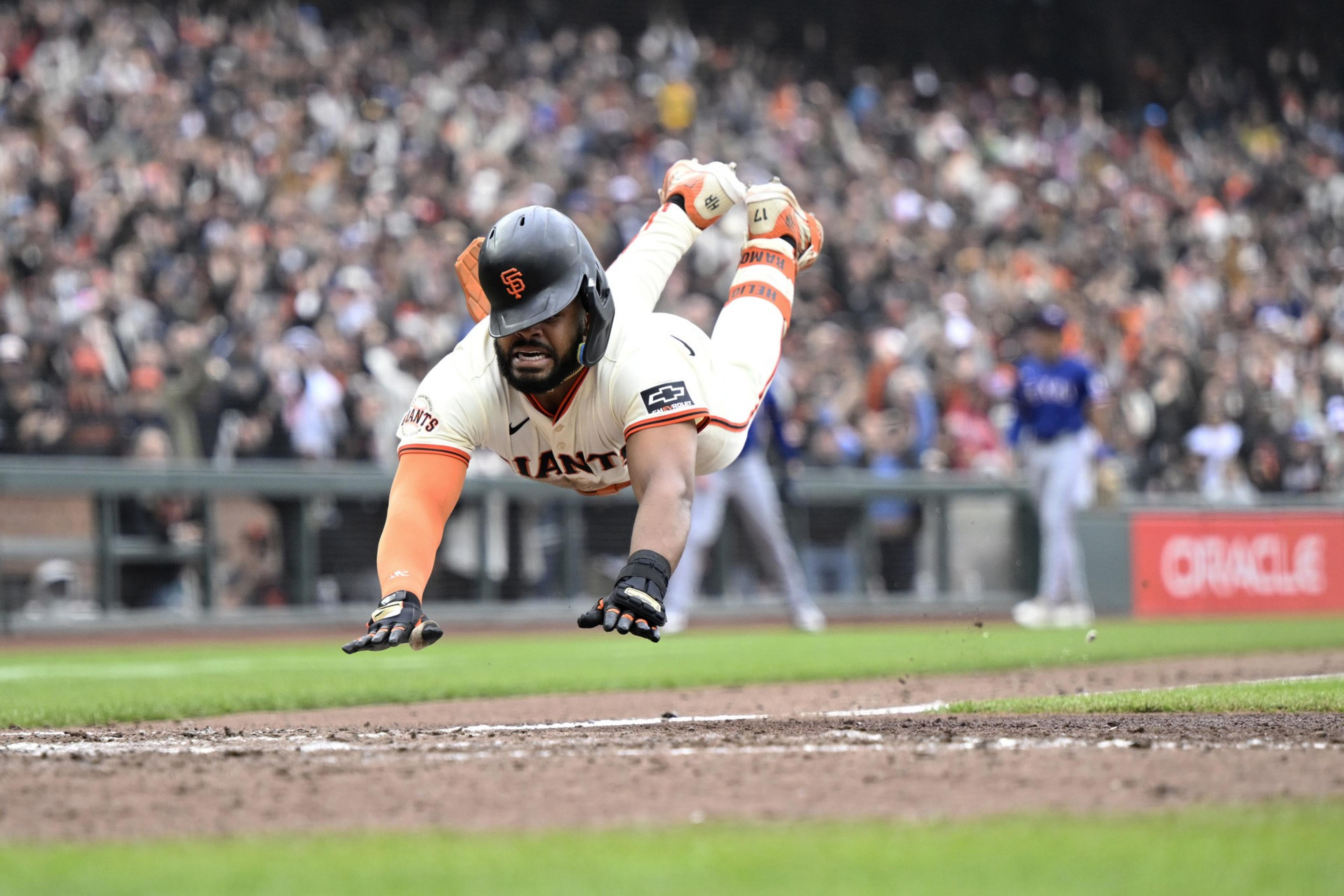 A baseball player in a Giants uniform is diving headfirst toward home plate. The crowd is blurred in the background as he extends his arms to score.