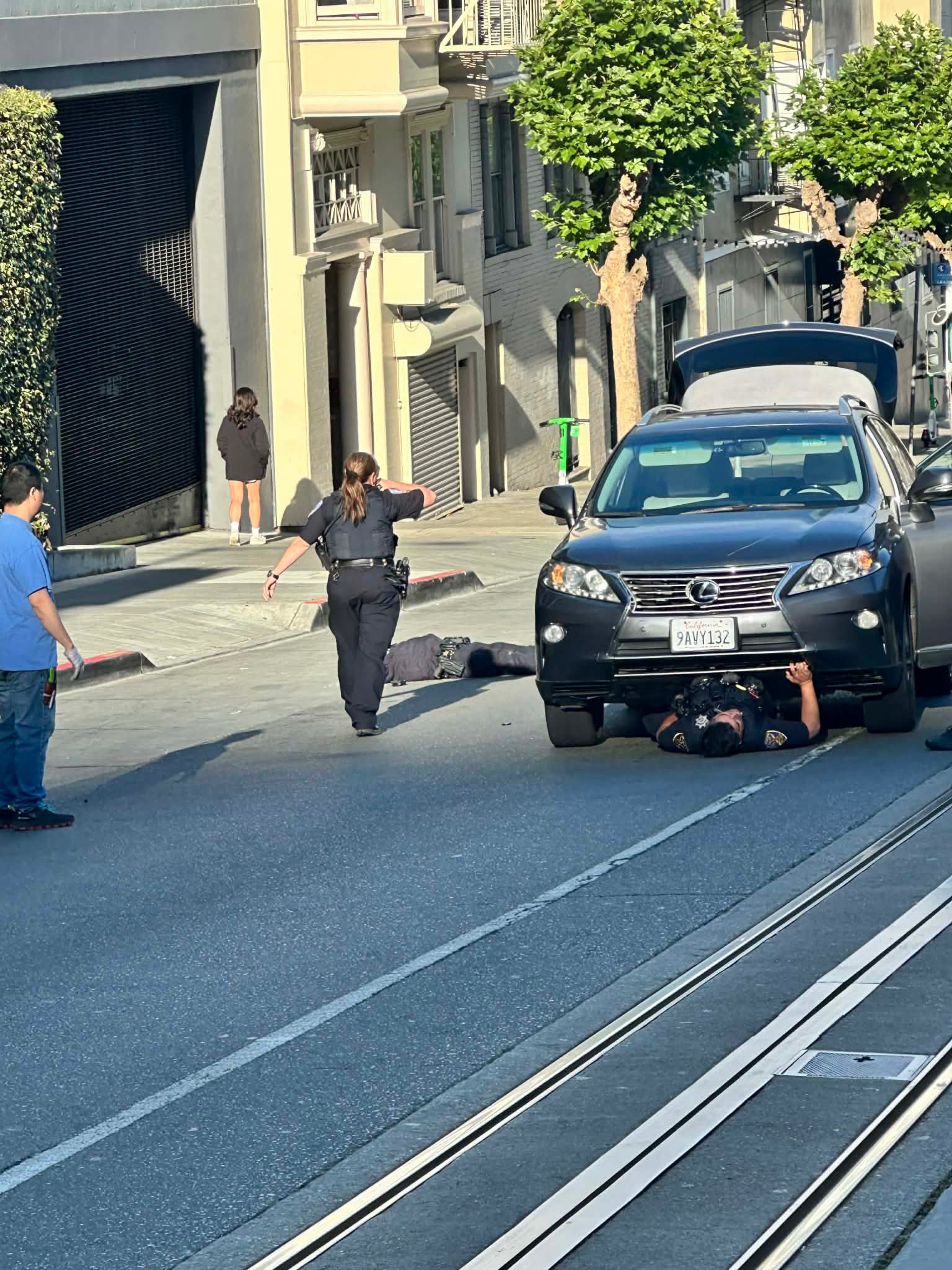 A police officer is lying under a black Lexus SUV performing a check, while another officer stands nearby and two people walk on the sidewalk.