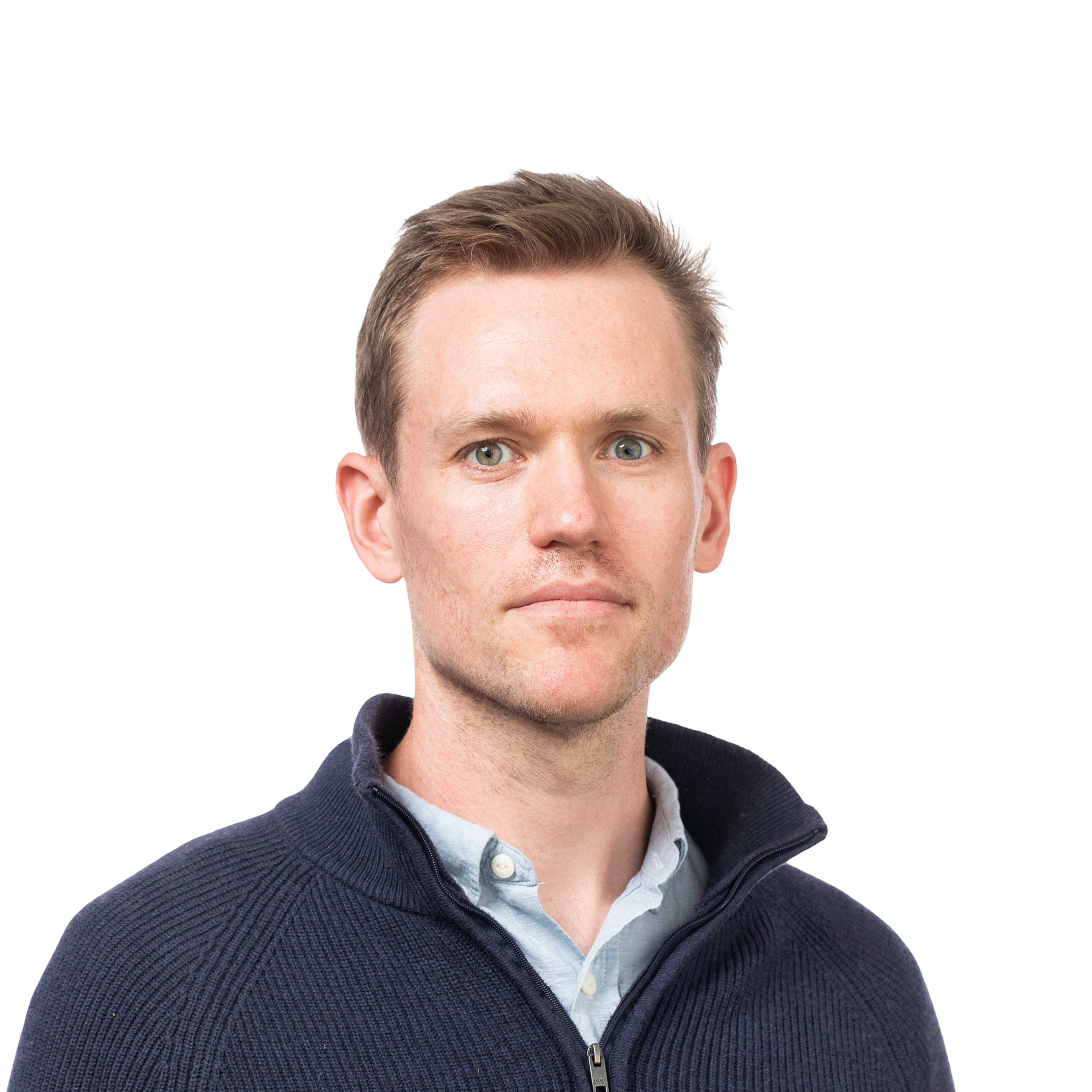A man with light skin, short light brown hair, and green eyes wears a navy zip-up sweater over a light blue collared shirt, looking straight ahead with a neutral expression.