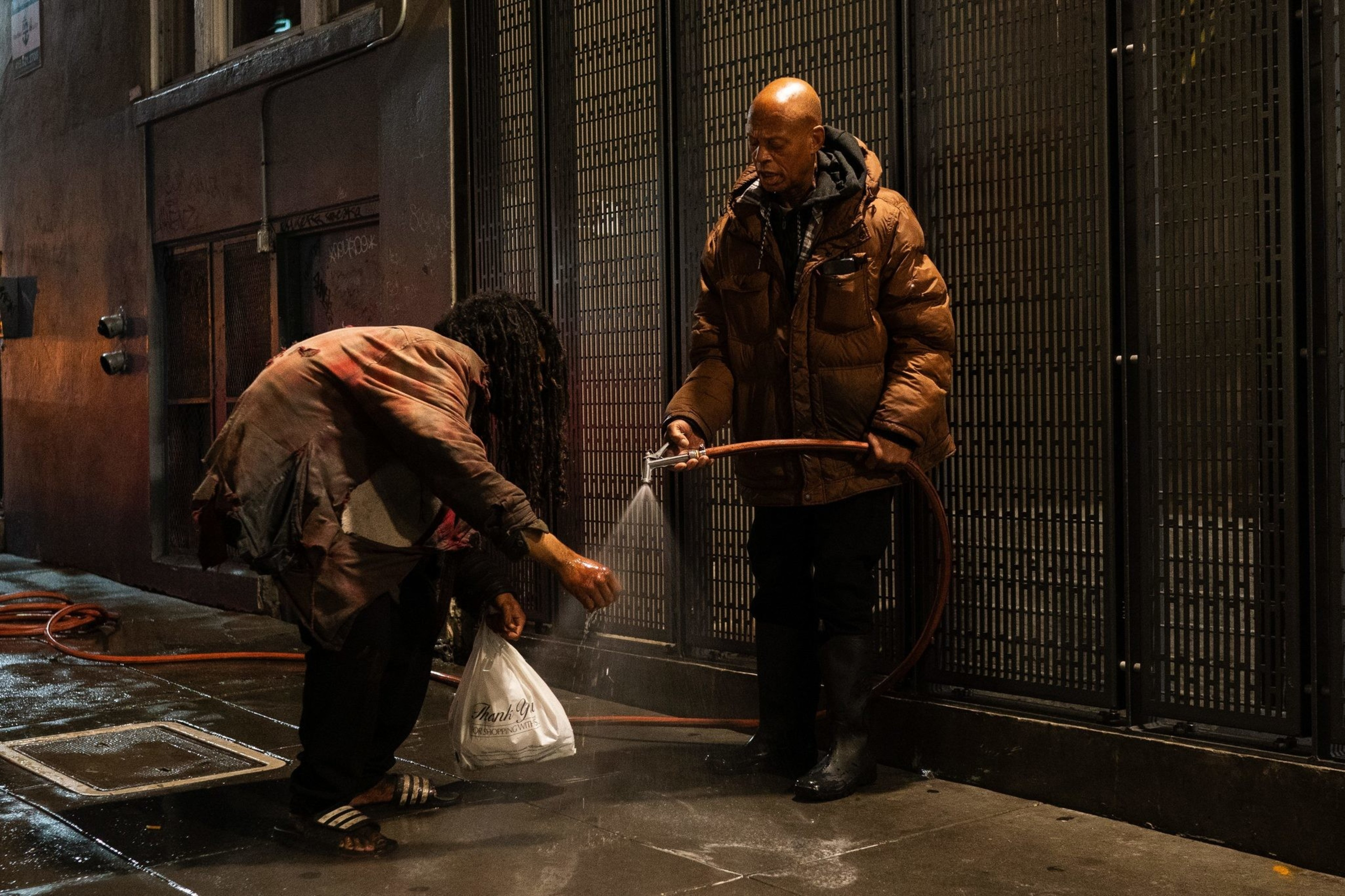 A man with a brown coat washes the hands of another person with a hose.