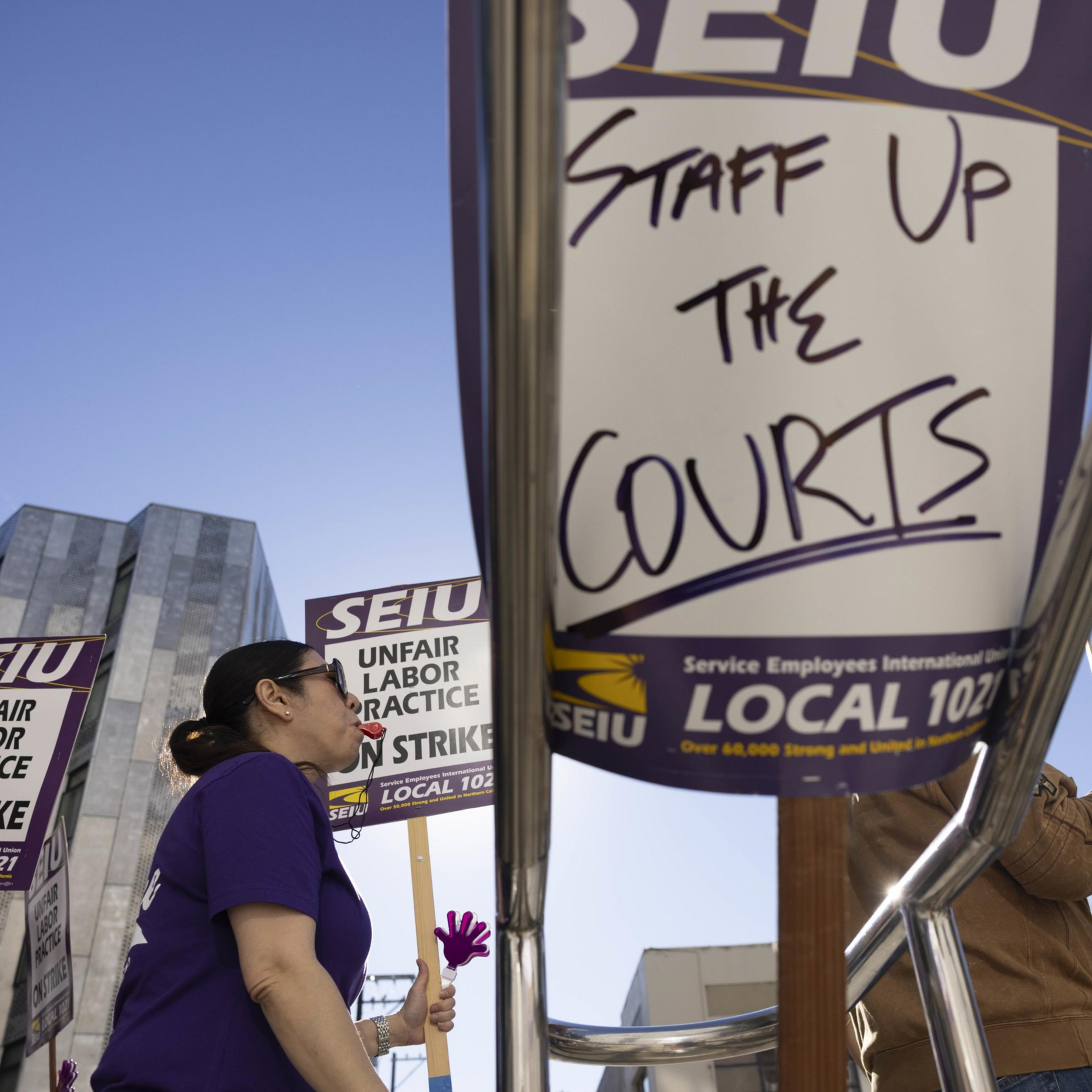 Protesters hold signs reading "SEIU Unfair Labor Practice" and "Staff Up The Courts." They stand in front of modern buildings under a clear blue sky.