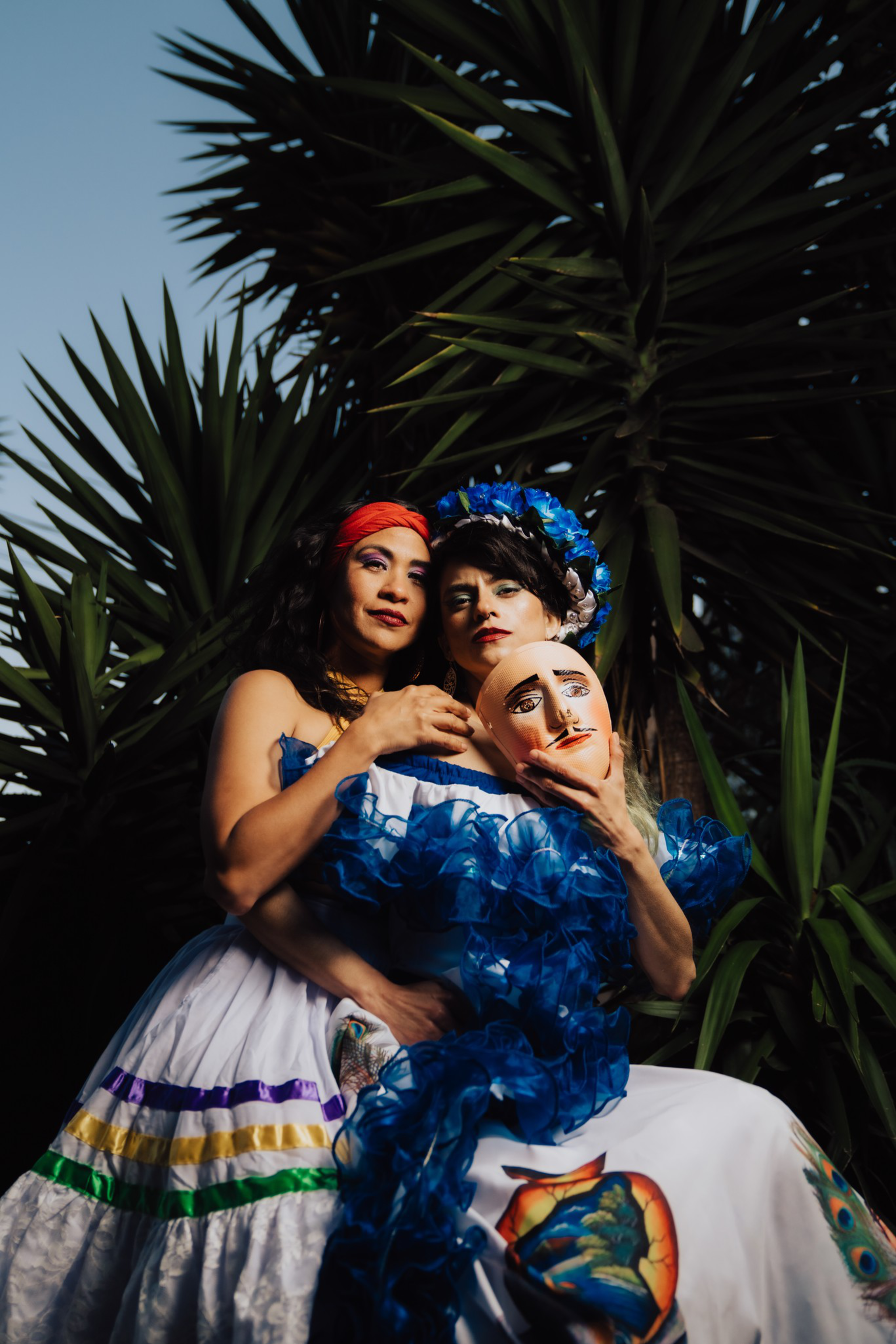 Two women in colorful, traditional dresses stand closely together, one holding a painted mask, surrounded by tall green plants under a dusky sky.