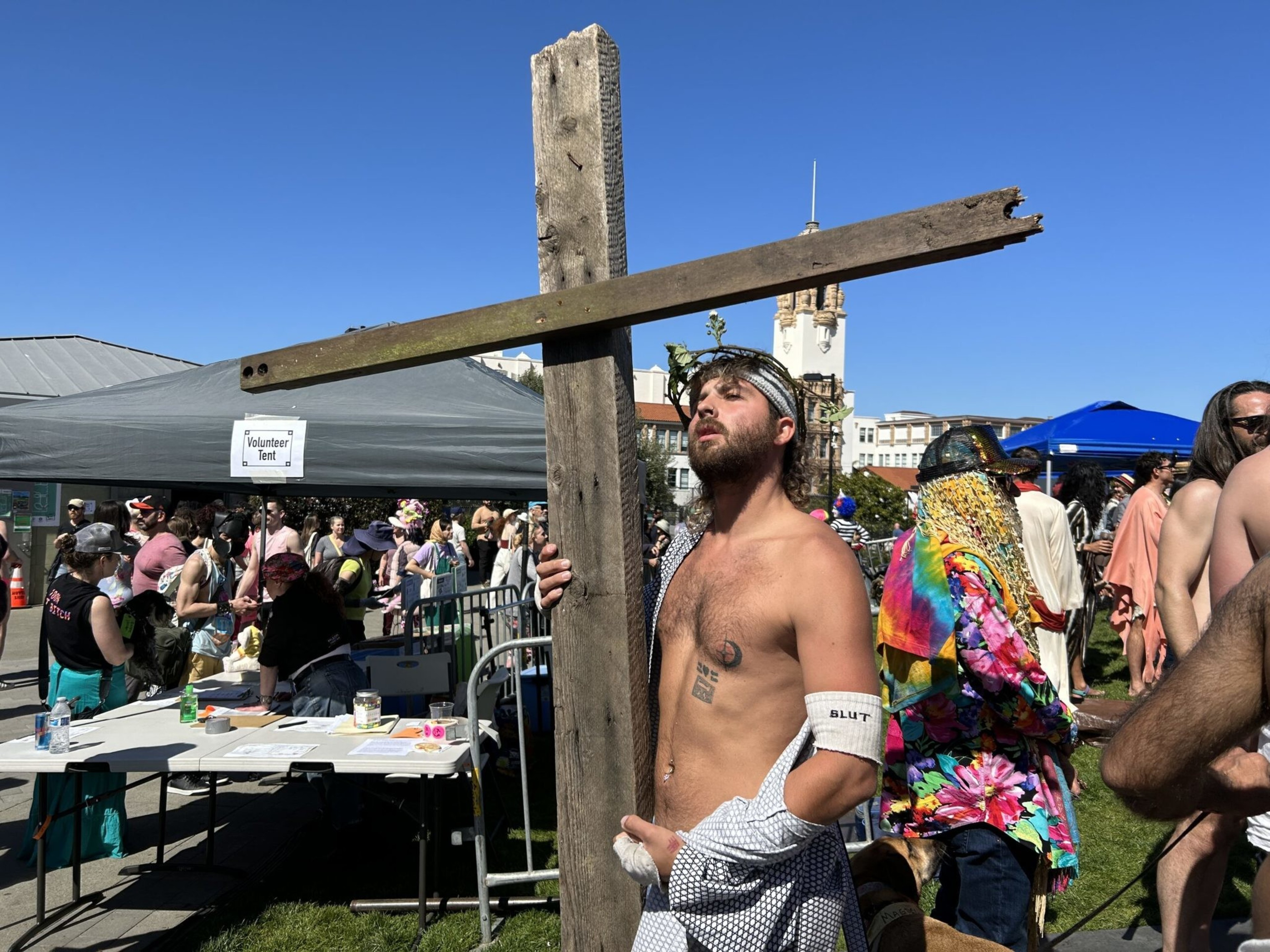 A shirtless man wearing a crown of thorns holds a large wooden cross in a crowded outdoor event under clear blue skies.