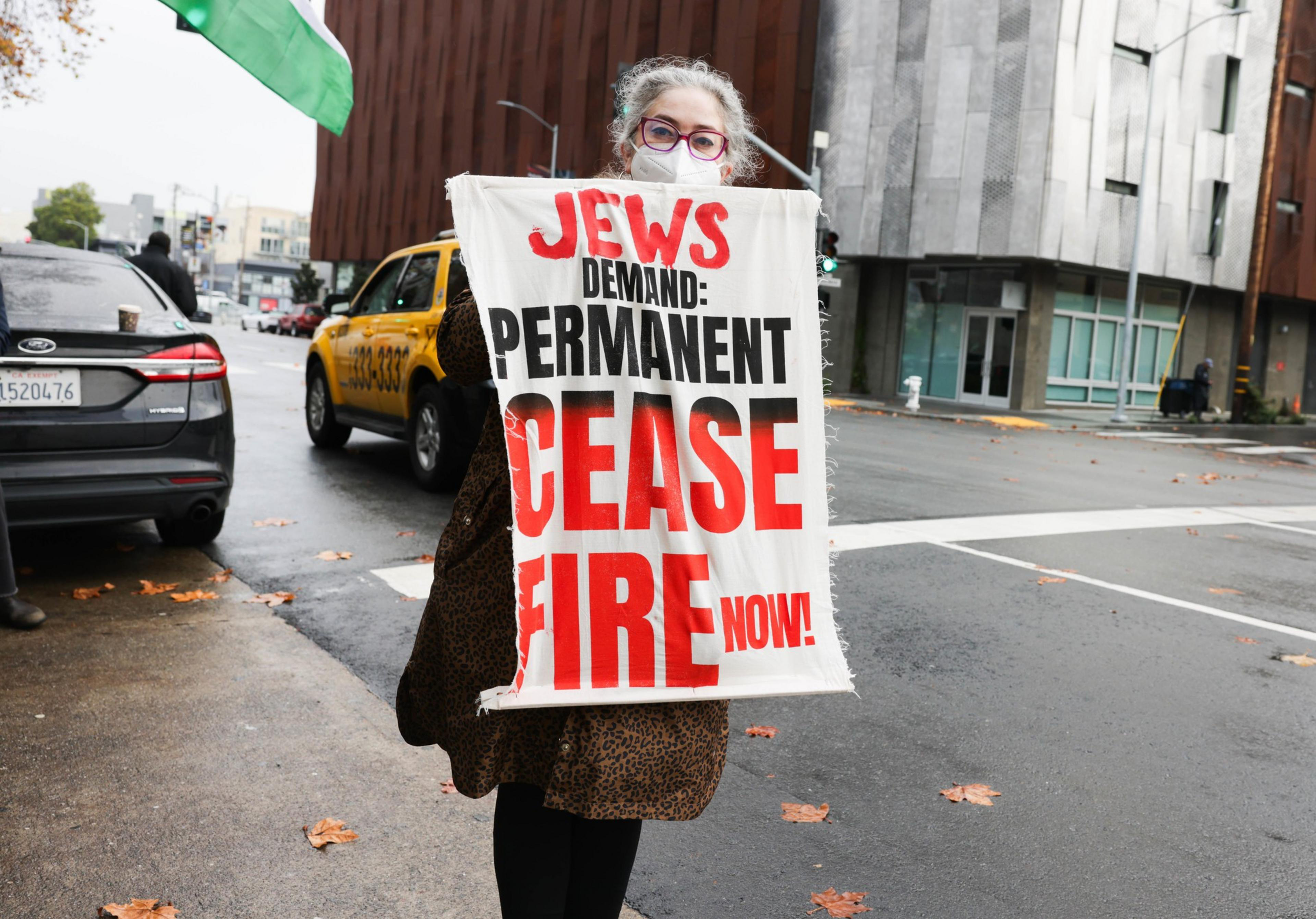 A person with white hair stands on the side walk with a sign. 