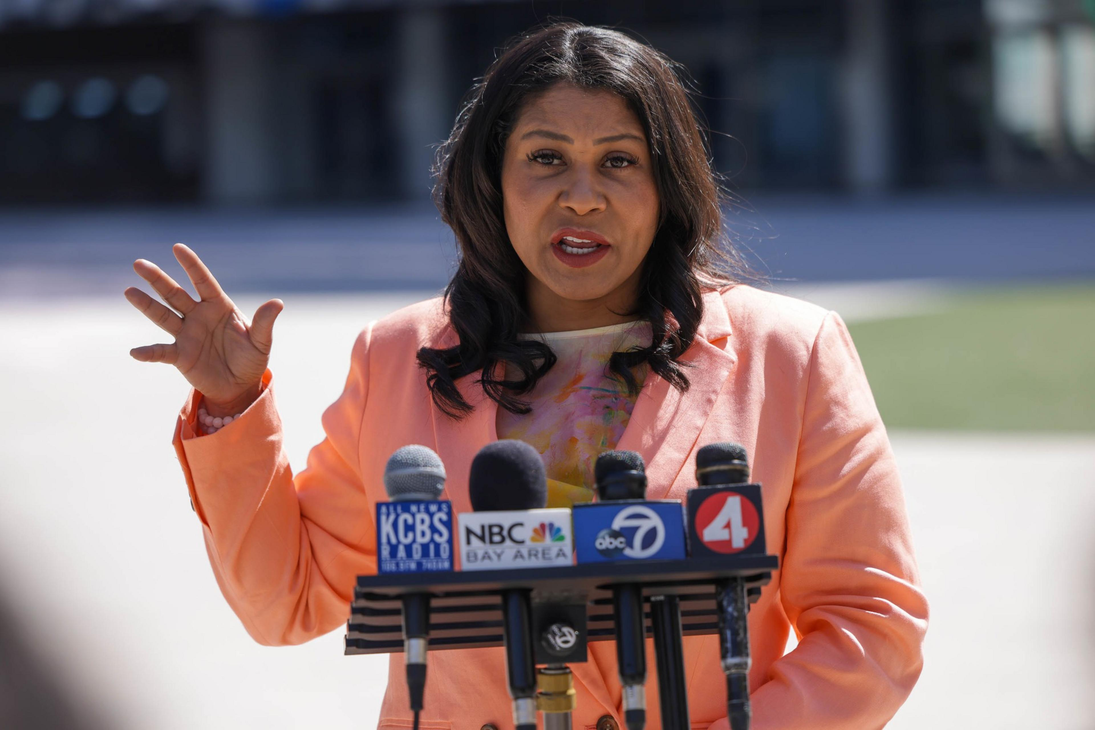A woman speaks at a podium outdoors, microphones in front, gesturing mid-speech.