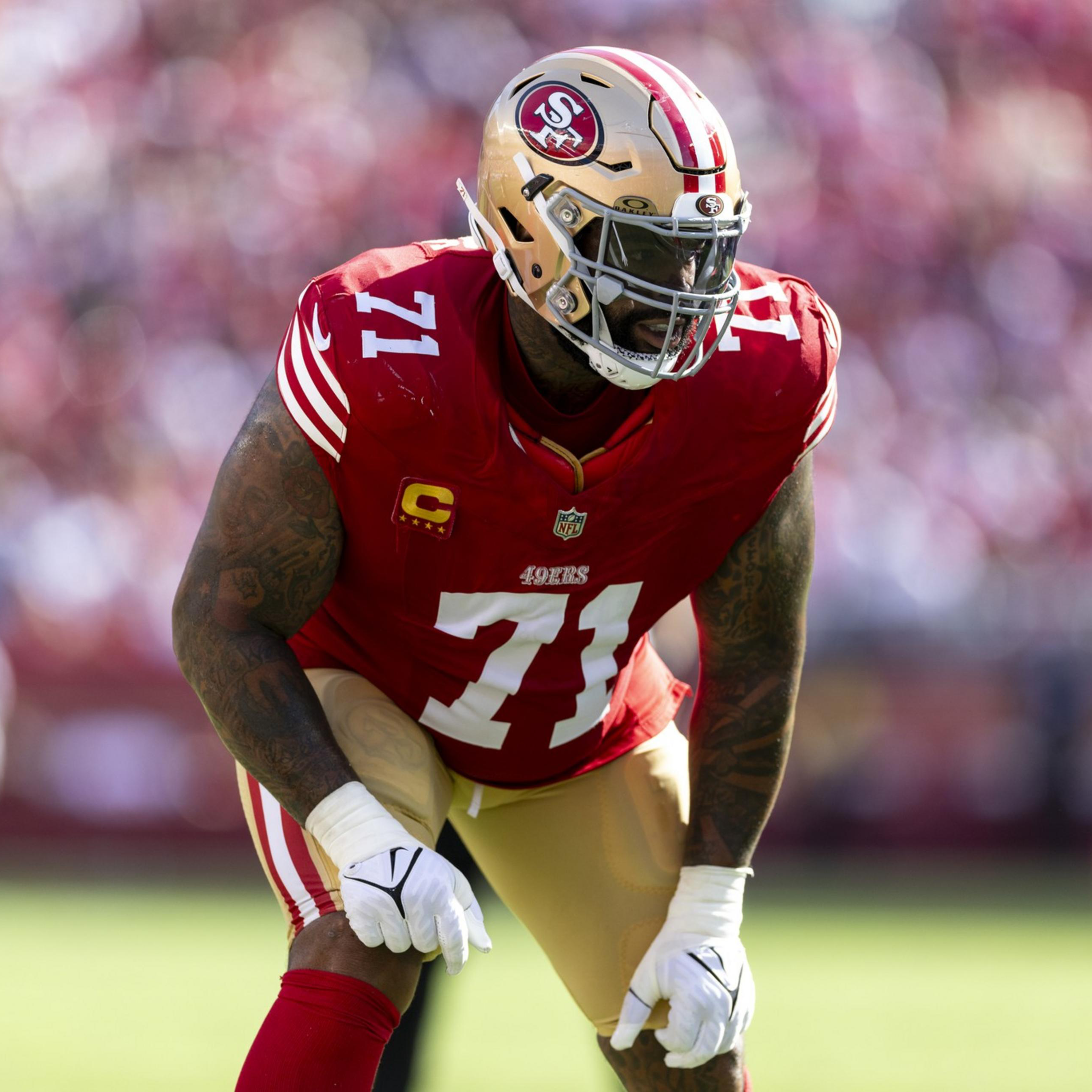 A San Francisco 49ers football player wearing a red jersey with number 71 and a gold helmet is poised in a defensive stance on a green field.