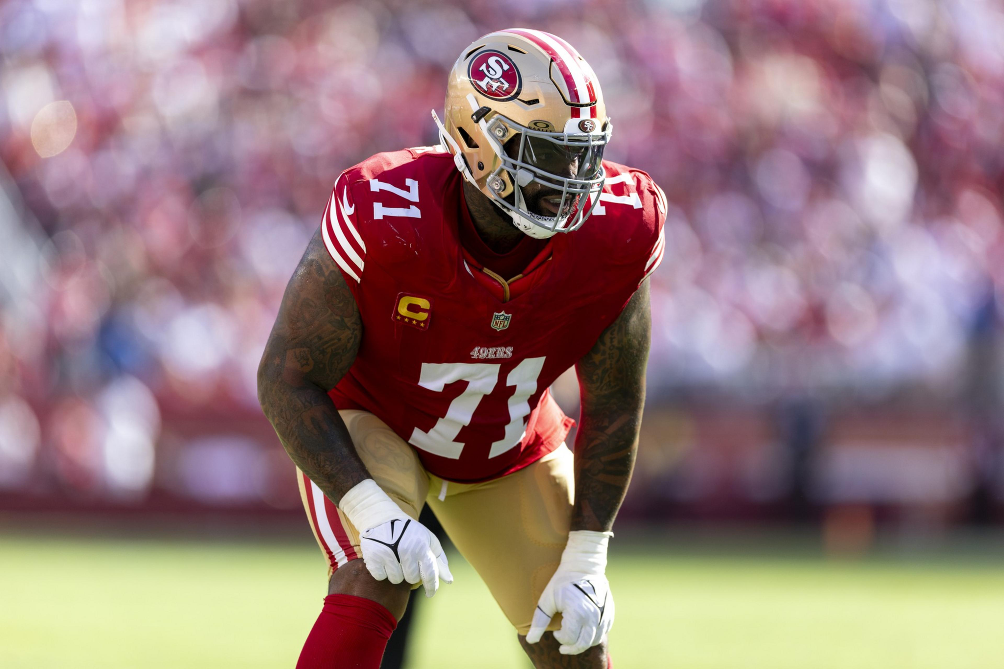 A San Francisco 49ers football player wearing a red jersey with number 71 and a gold helmet is poised in a defensive stance on a green field.