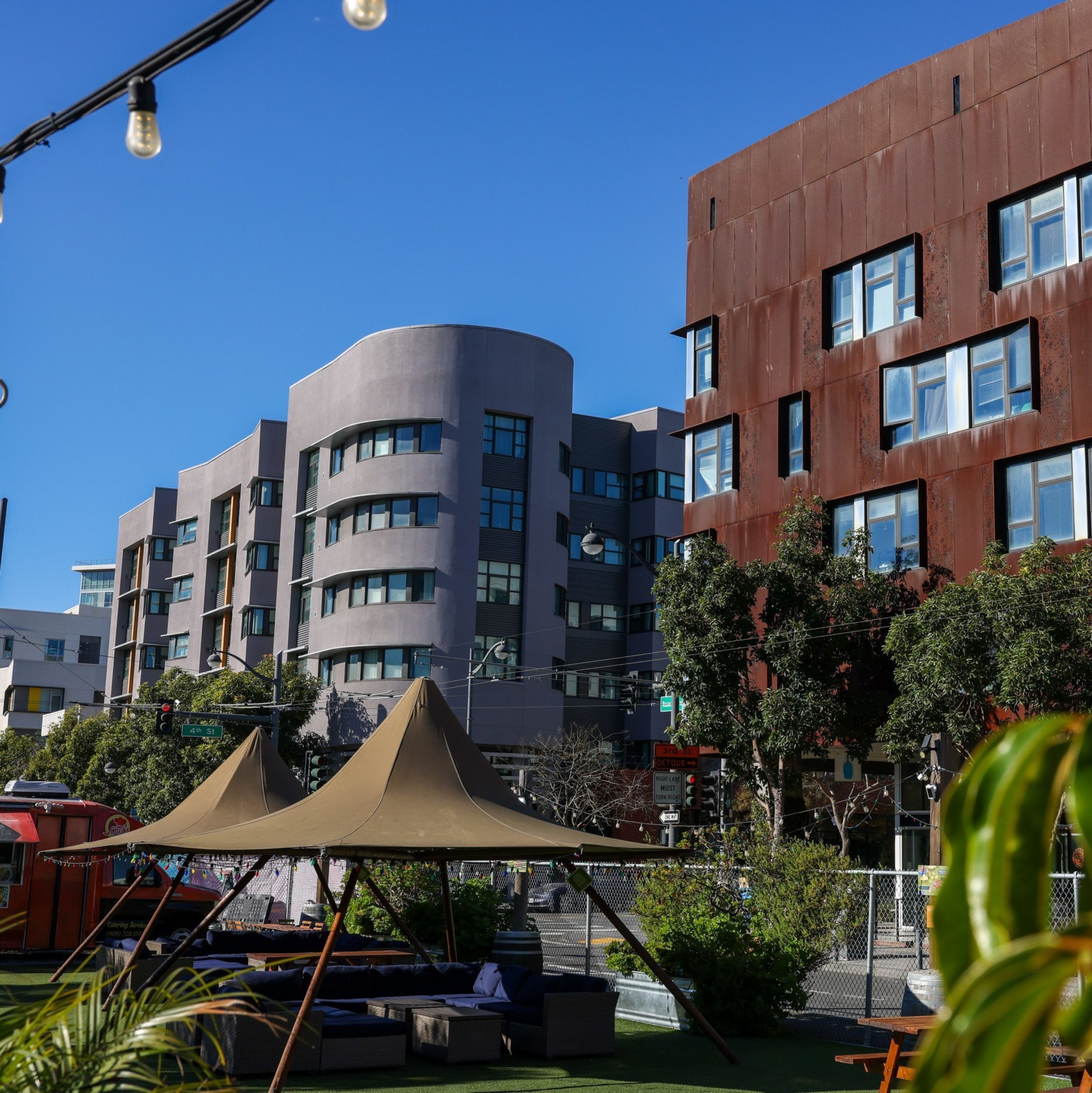A modern urban scene features two distinctive buildings, one grey and curved, the other rust-colored. Trees, plants, and string lights add a touch of greenery.