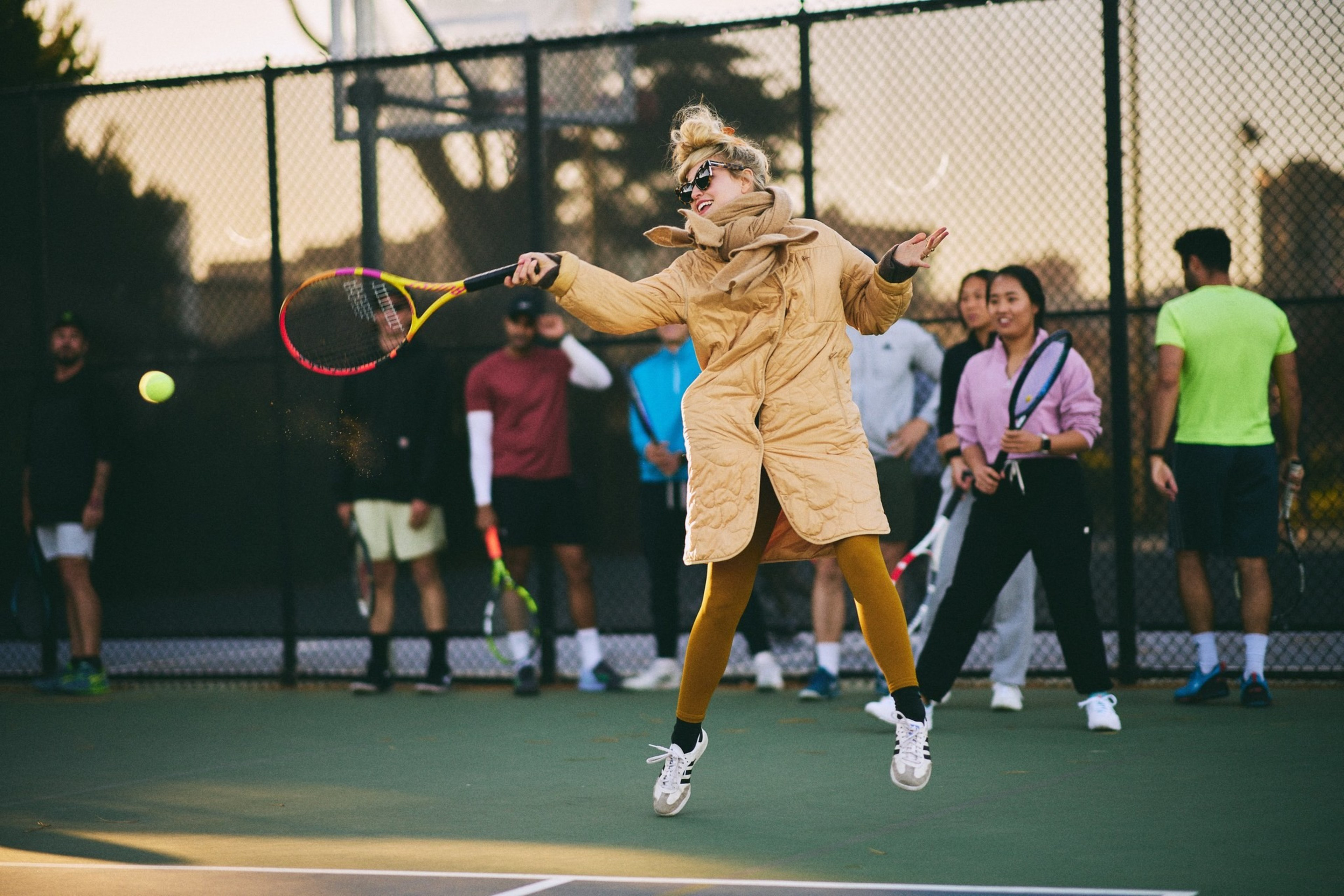 A person in a long coat and sunglasses is jumping to hit a tennis ball on a court, surrounded by seven spectators casually dressed, holding rackets.