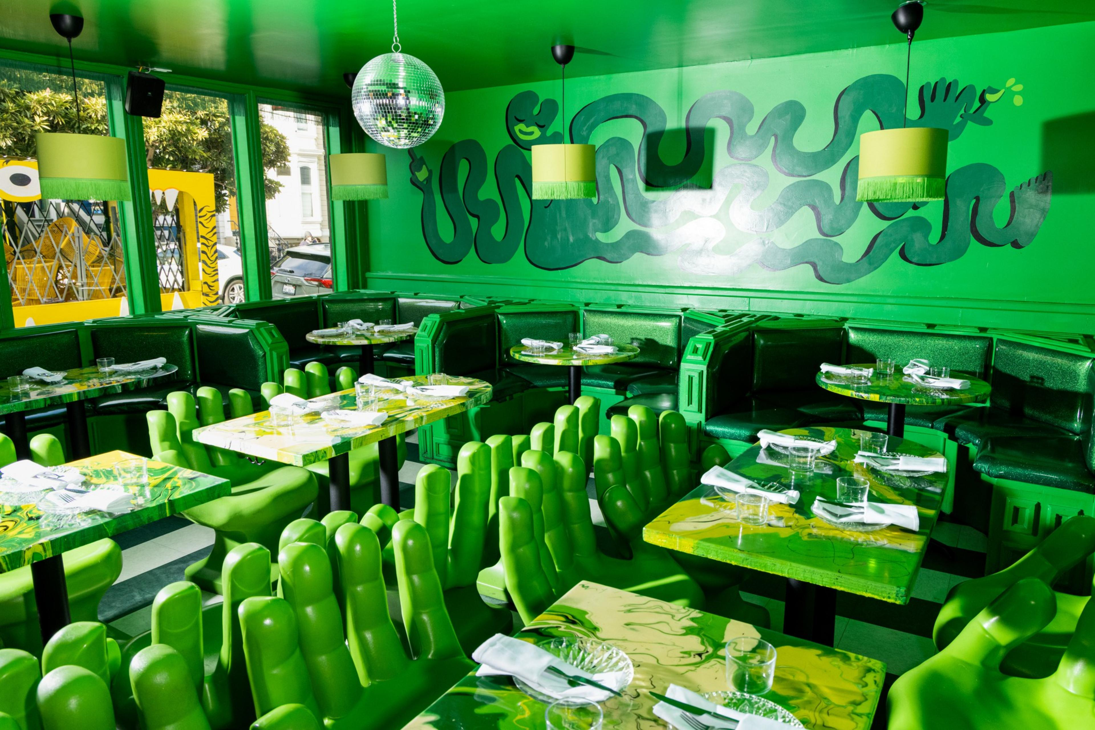 A vibrant green dining room features hand-shaped chairs, marble-patterned tables, green pendant lights, a disco ball, and snake-themed wall art.