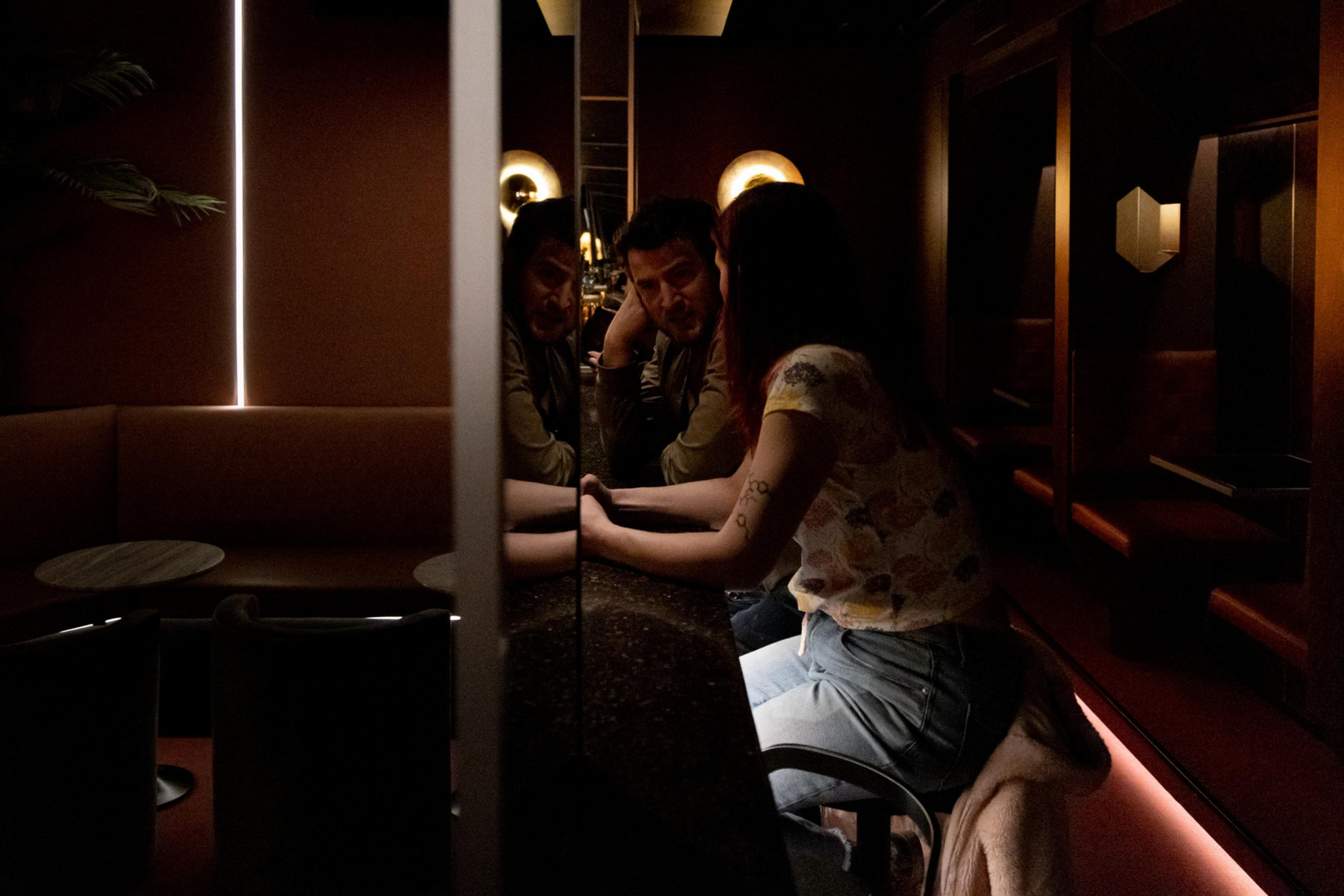 Two people sit close at a dark bar, their hands touching across the counter, with a dimly lit, cozy atmosphere and warm wall lights behind them.