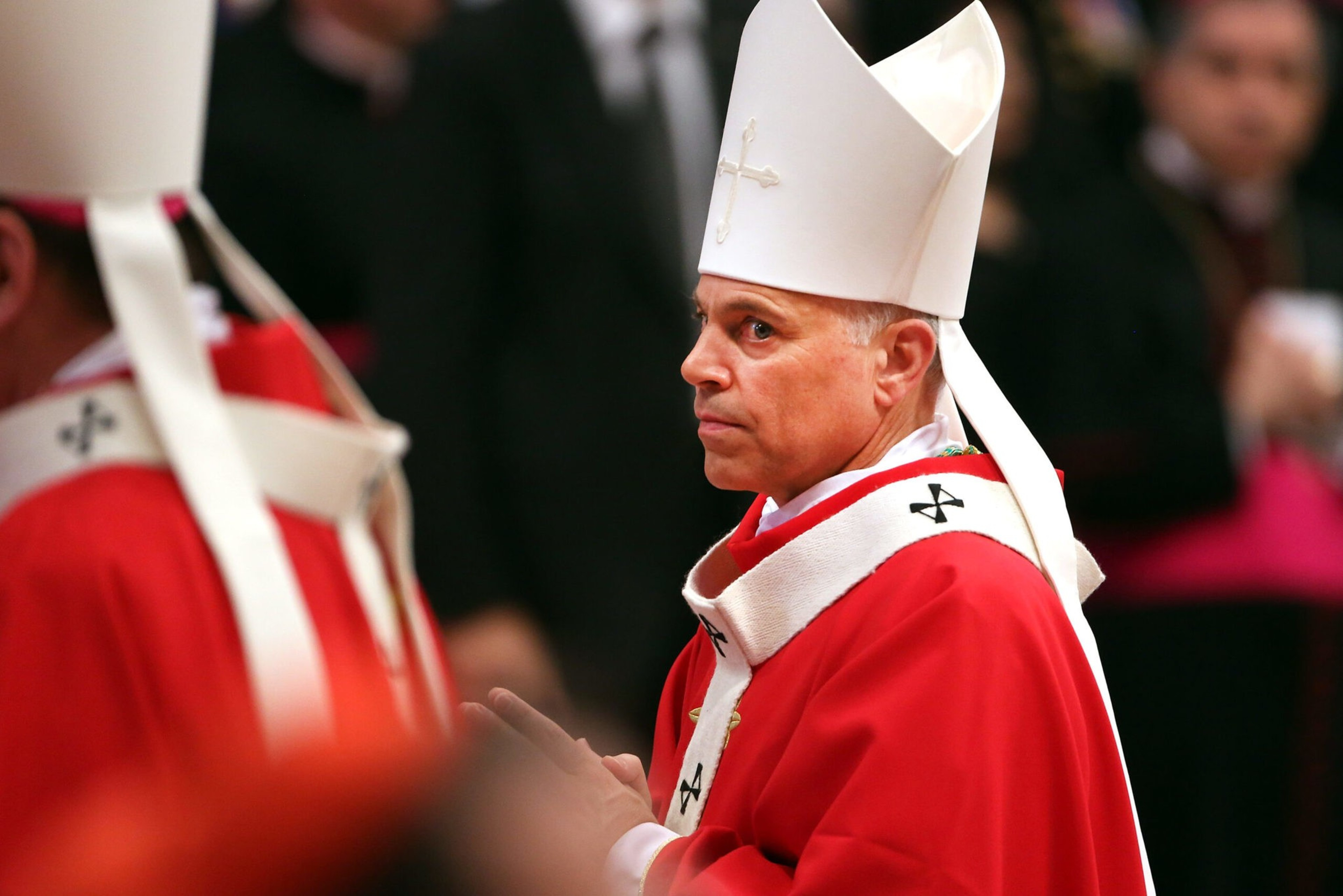A clergyman in a red robe with white trim and a white mitre hat with a cross, standing solemnly in a church setting.
