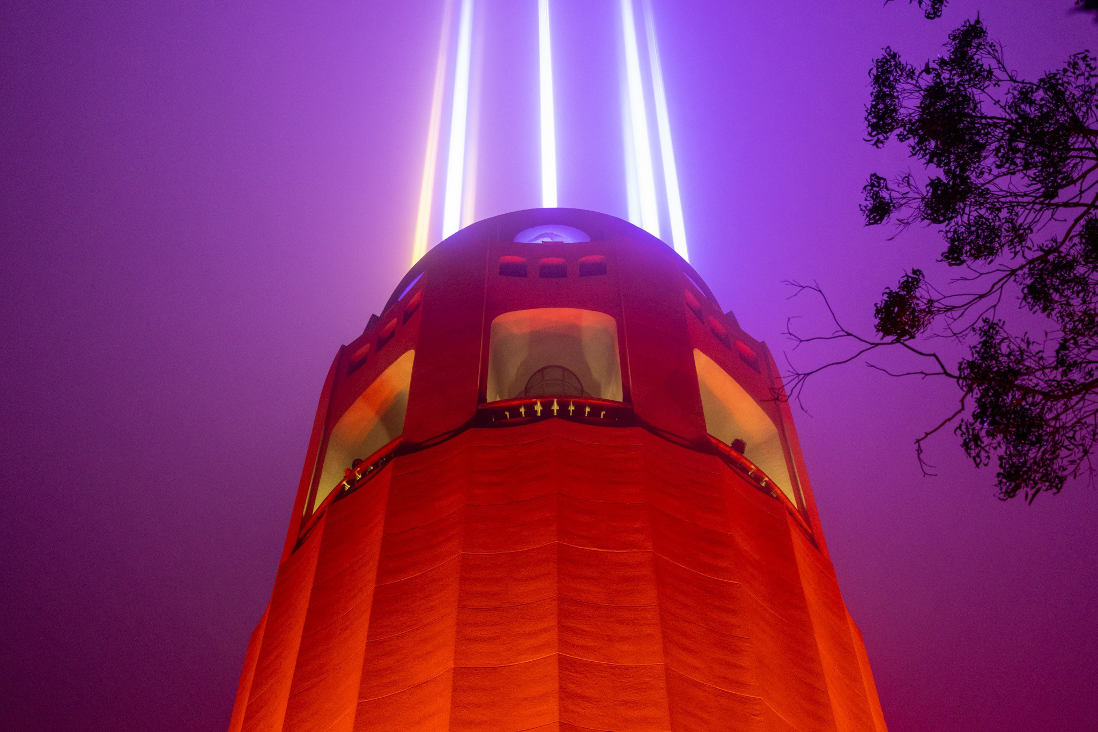A tall, cylindrical red tower emits three vertical beams of bright white and purple light into a foggy purple sky, framed by tree branches on the right.