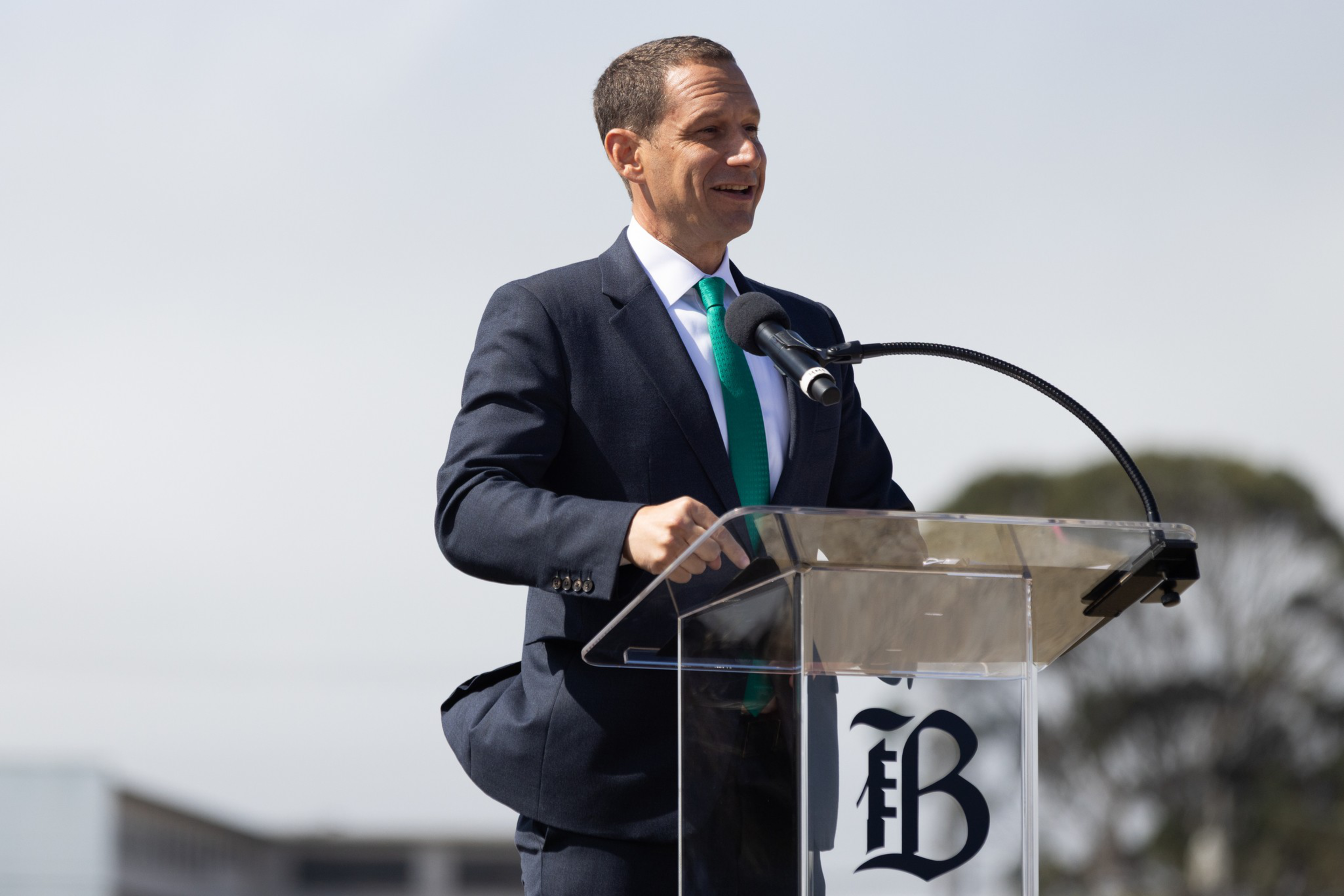 A man in a dark suit and green tie speaks smiling at a clear podium with a microphone against a blurry outdoor background.