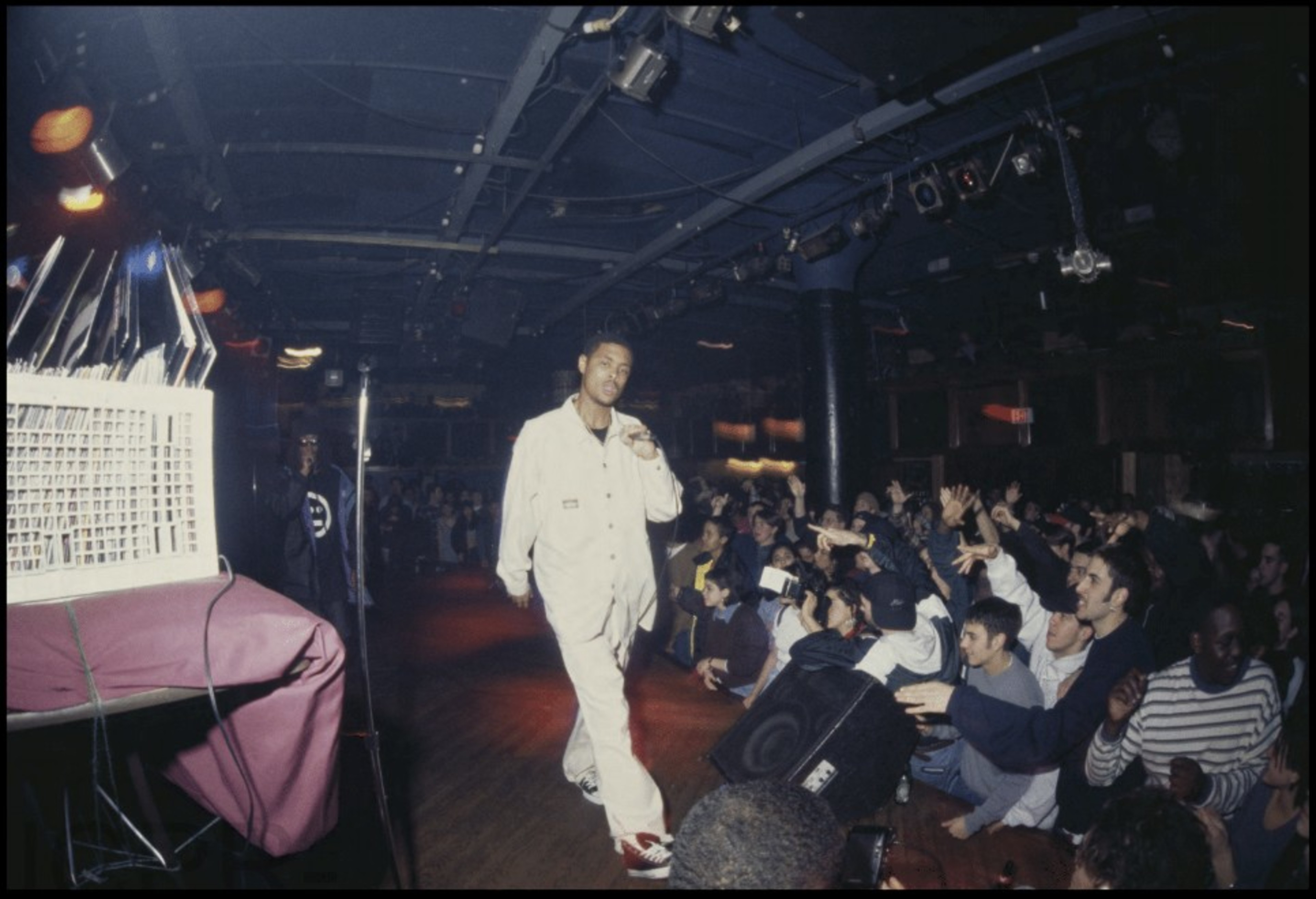 A person in a white outfit is performing on stage with a crowd enthusiastically cheering. The setting is dimly lit, resembling a concert or nightclub.