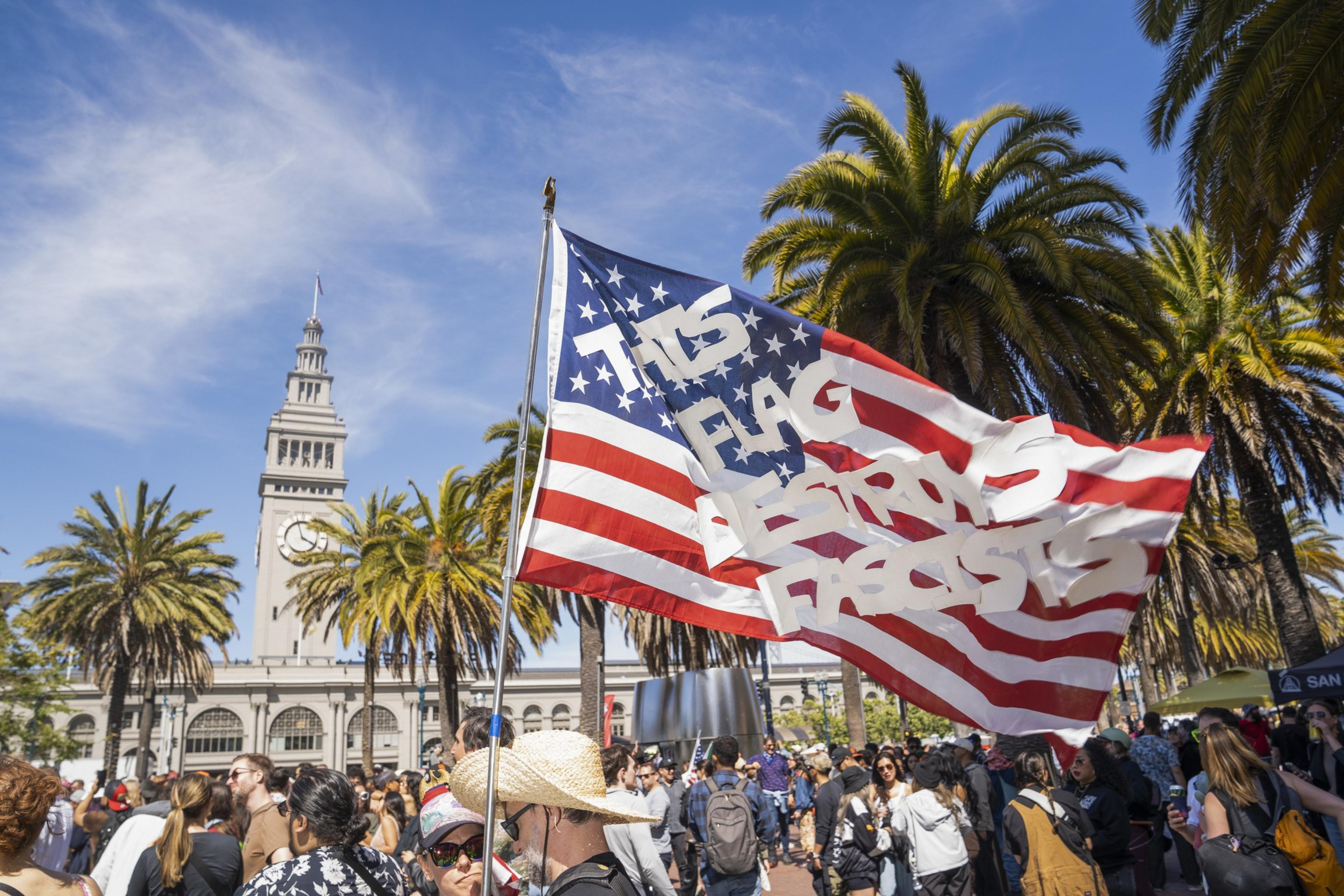 A crowd gathers near palm trees and a clock tower. An American flag with the text &quot;This flag destroys fascists&quot; is prominently displayed.