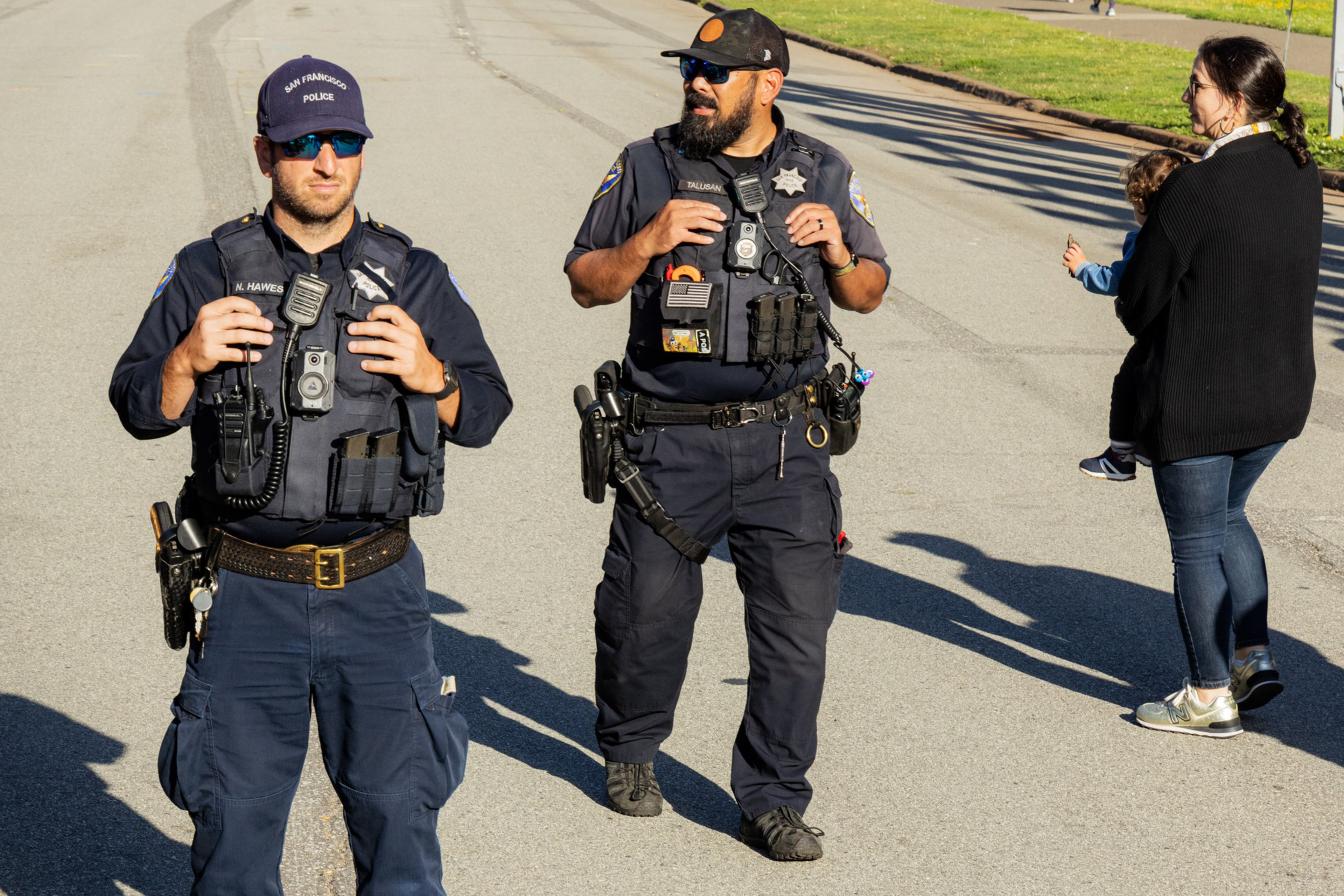Two police officers in uniform stand on a street, with one holding a radio. A woman holding a child is nearby, with grass visible in the background.