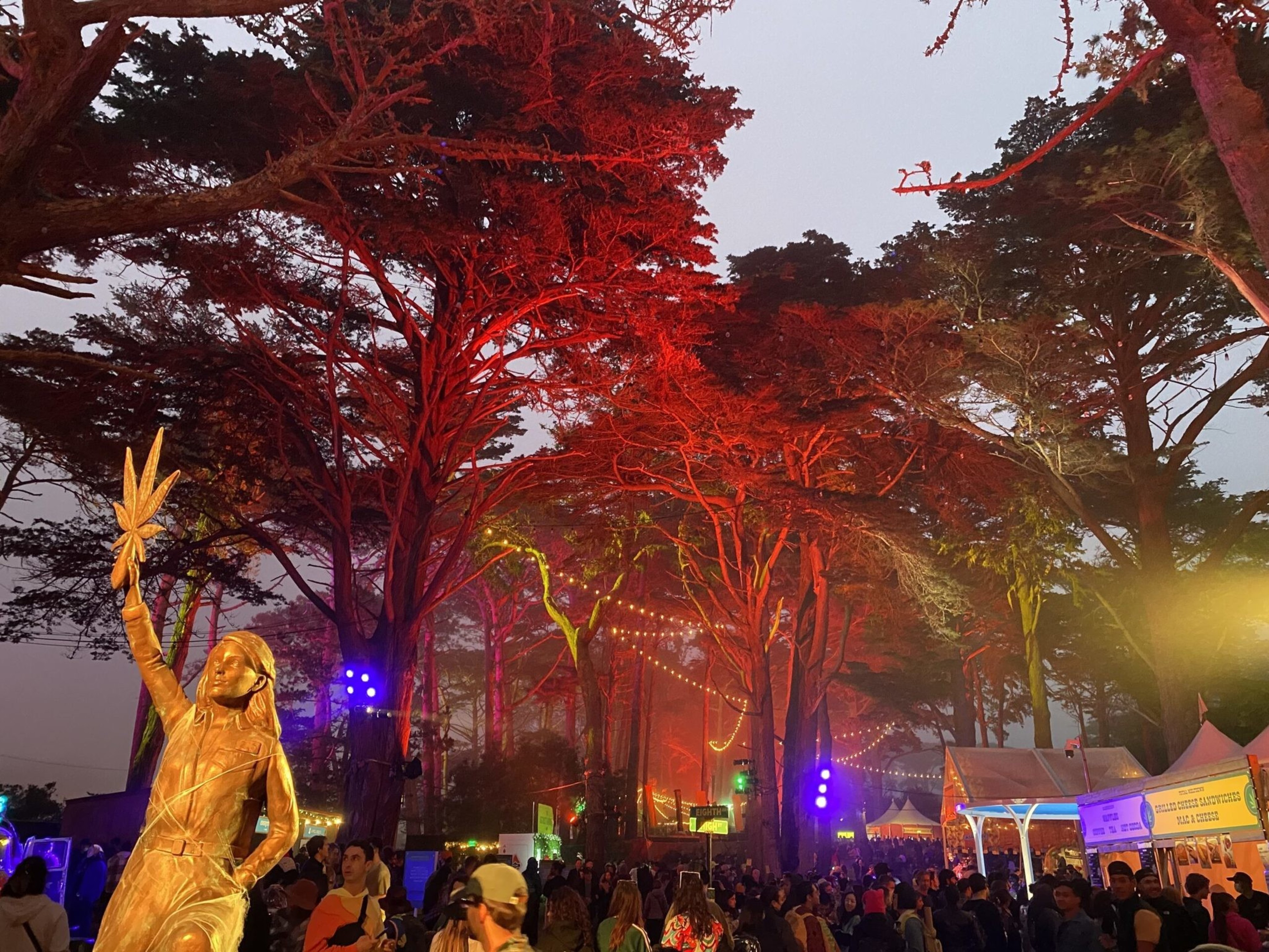 A golden statue of a person holding a leaf stands among tall trees lit with red lights, while a crowd gathers near colorful festival tents at dusk.