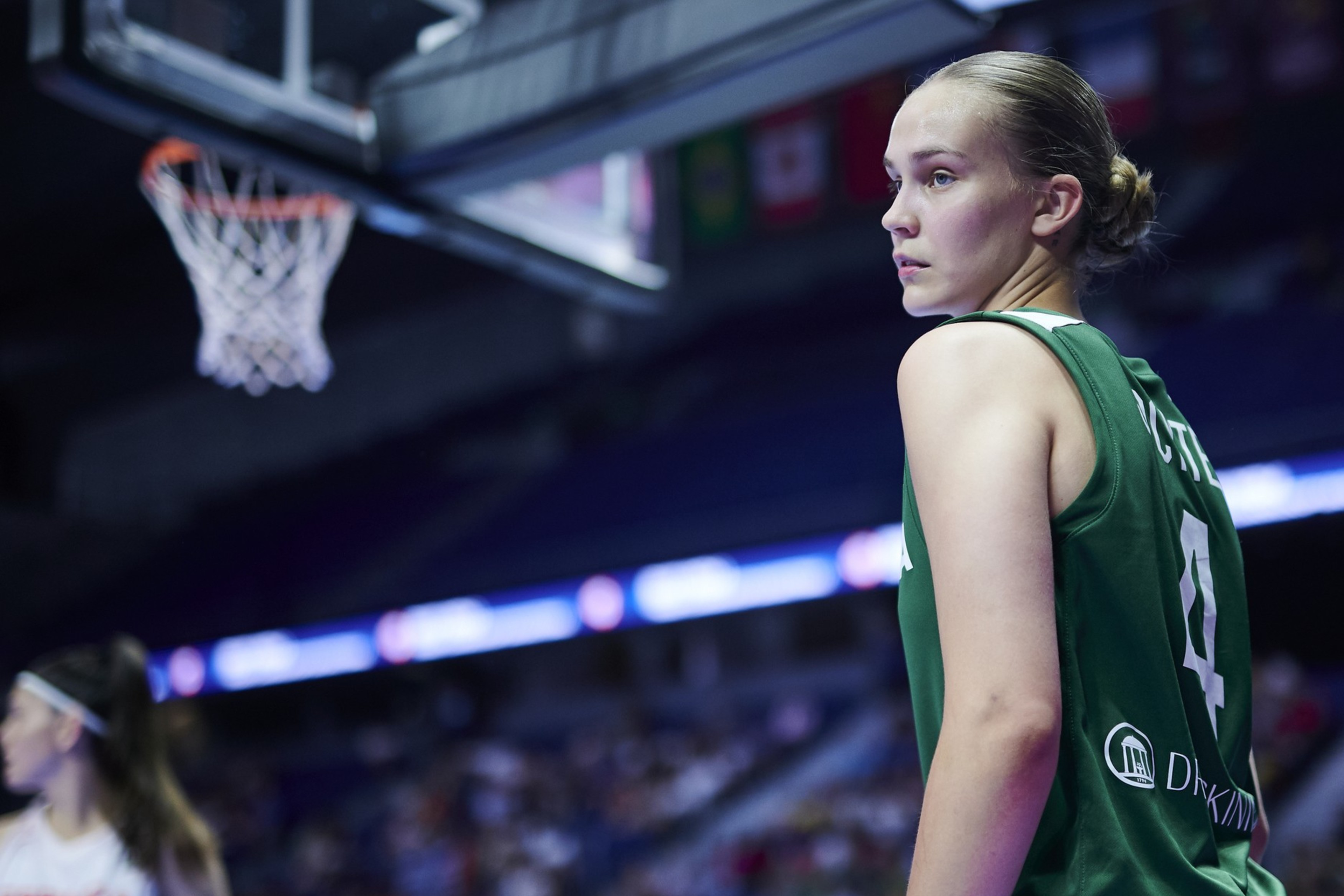 A basketball player in a green jersey looks over her shoulder on the court, with a blurred basketball hoop above and a crowded stadium in the background.