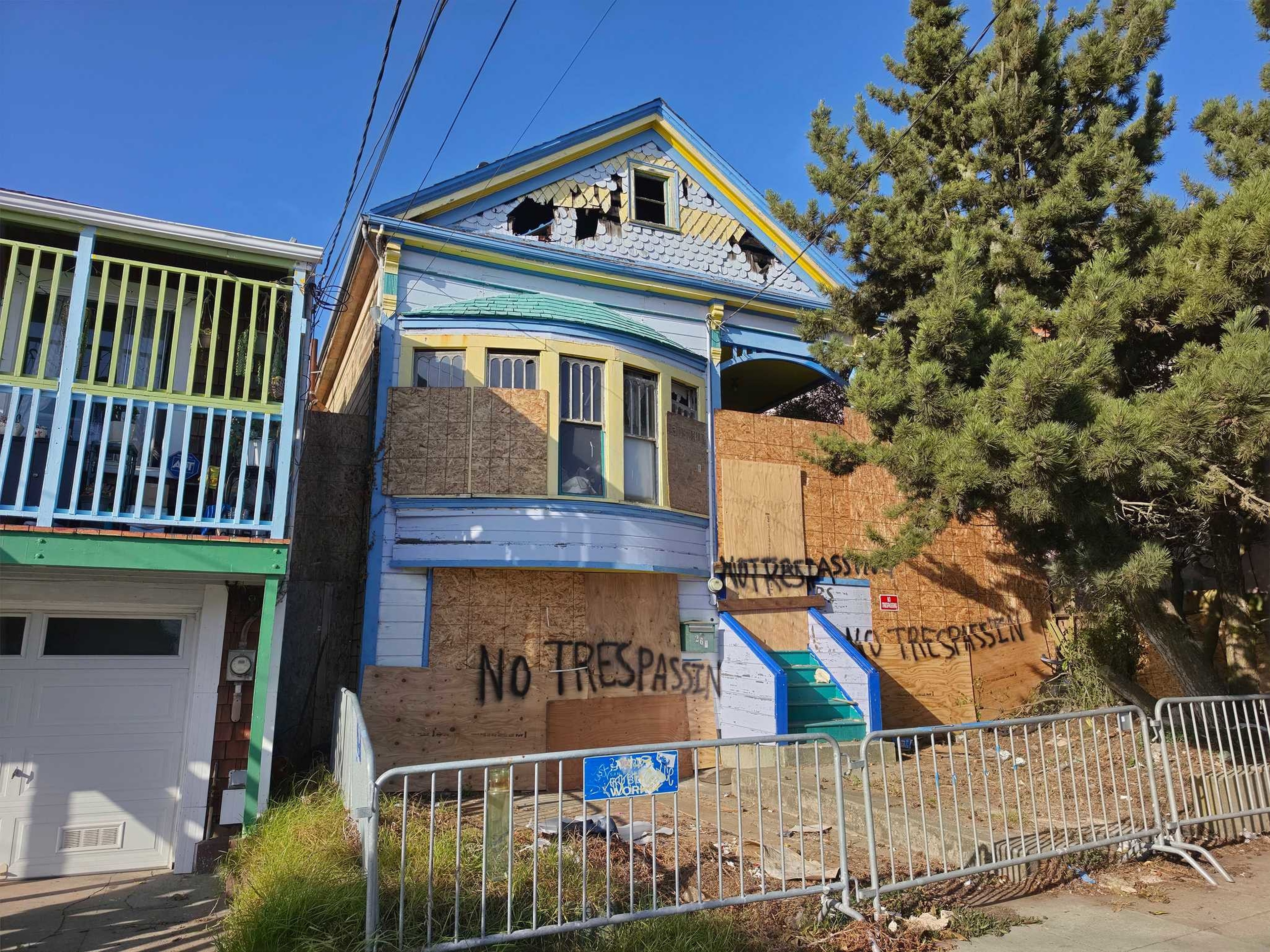 A boarded-up, damaged house with "NO TRESPASSIN" spray-painted on the plywood, metal barricades in front, and a large tree on the right side.