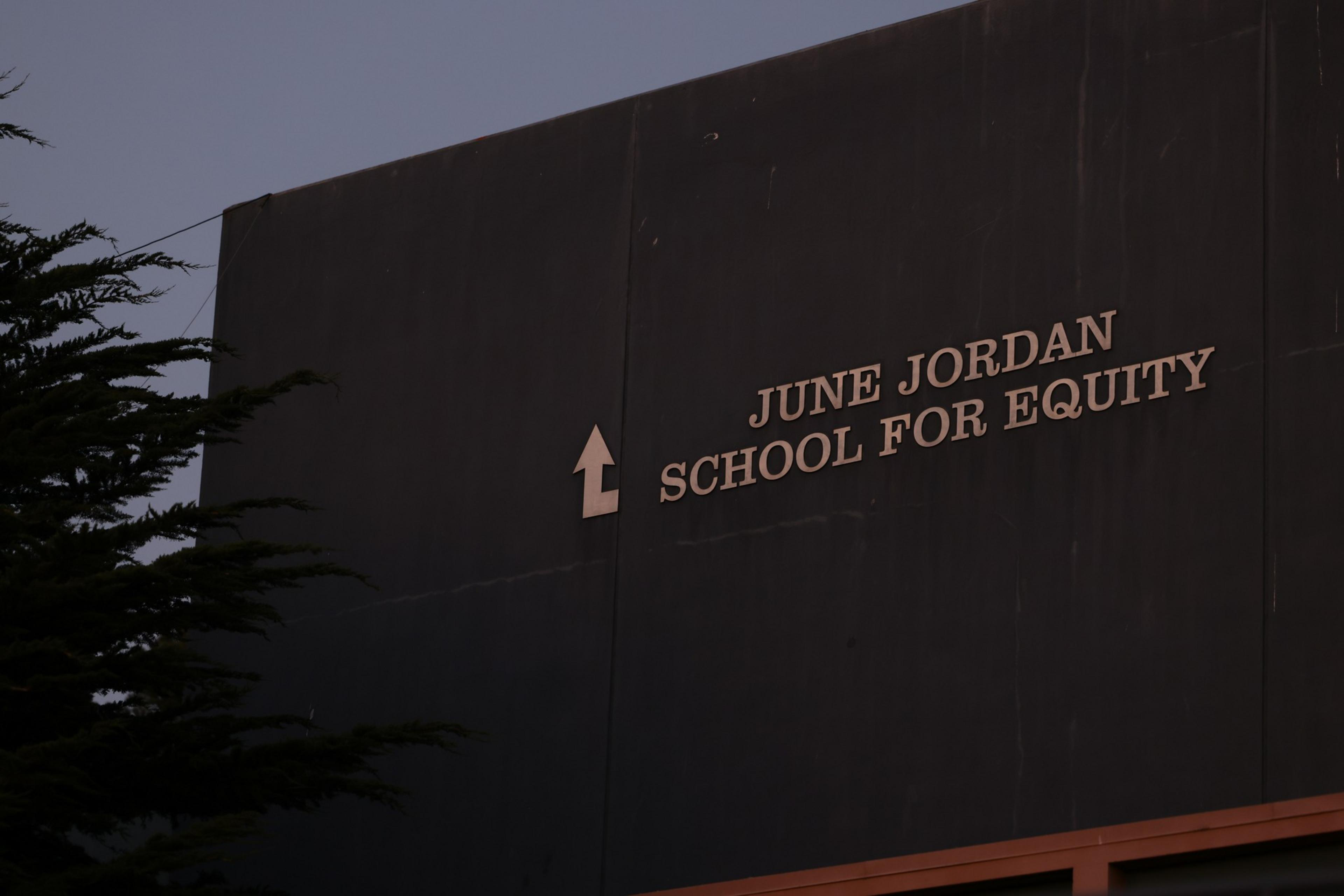 "June Jordan School for Equity" is written on a dark building wall with a left-turn arrow next to the text, and some tree branches are visible.