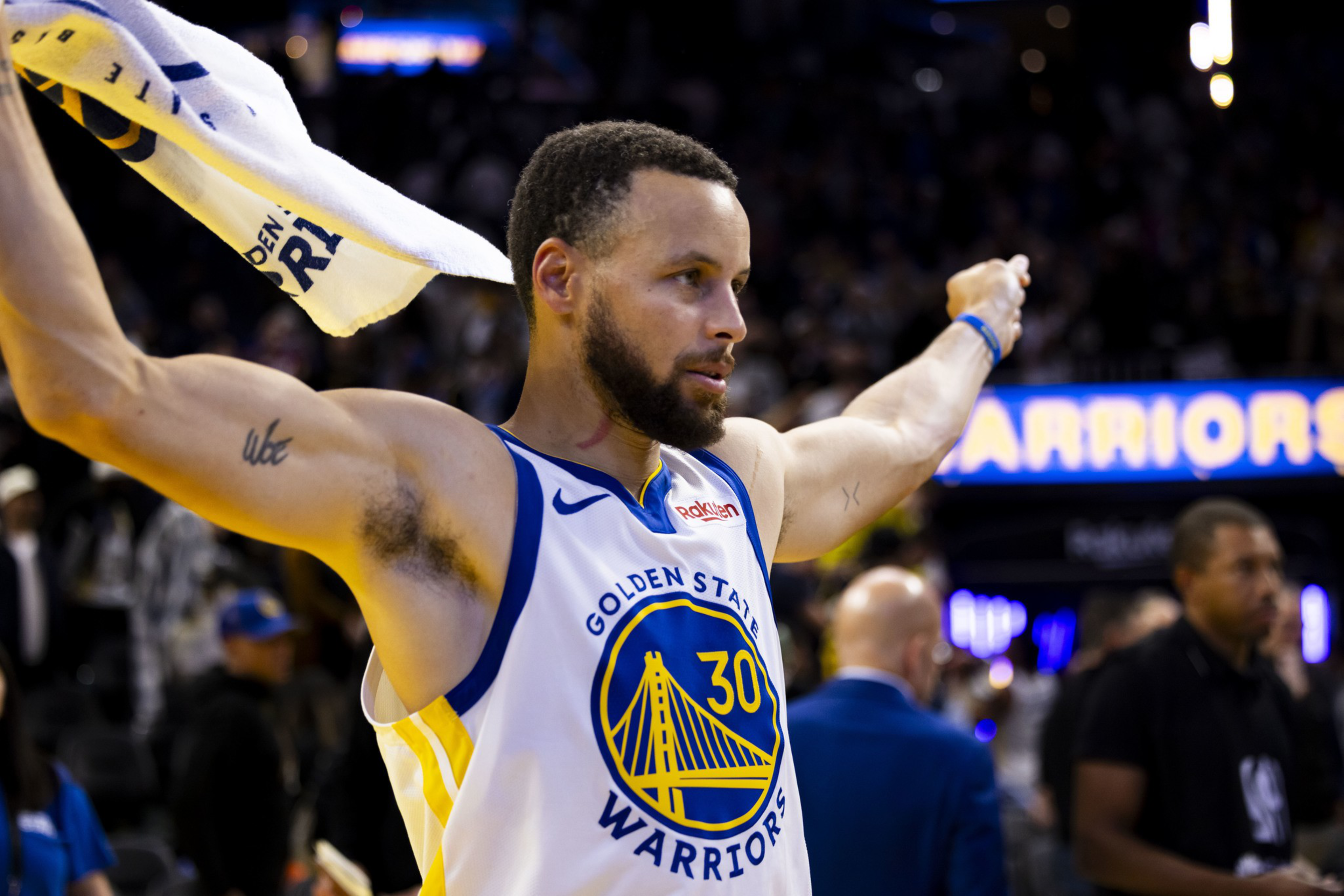 A basketball player wearing a Golden State Warriors jersey with number 30 holds a towel overhead, celebrating with arms outstretched.