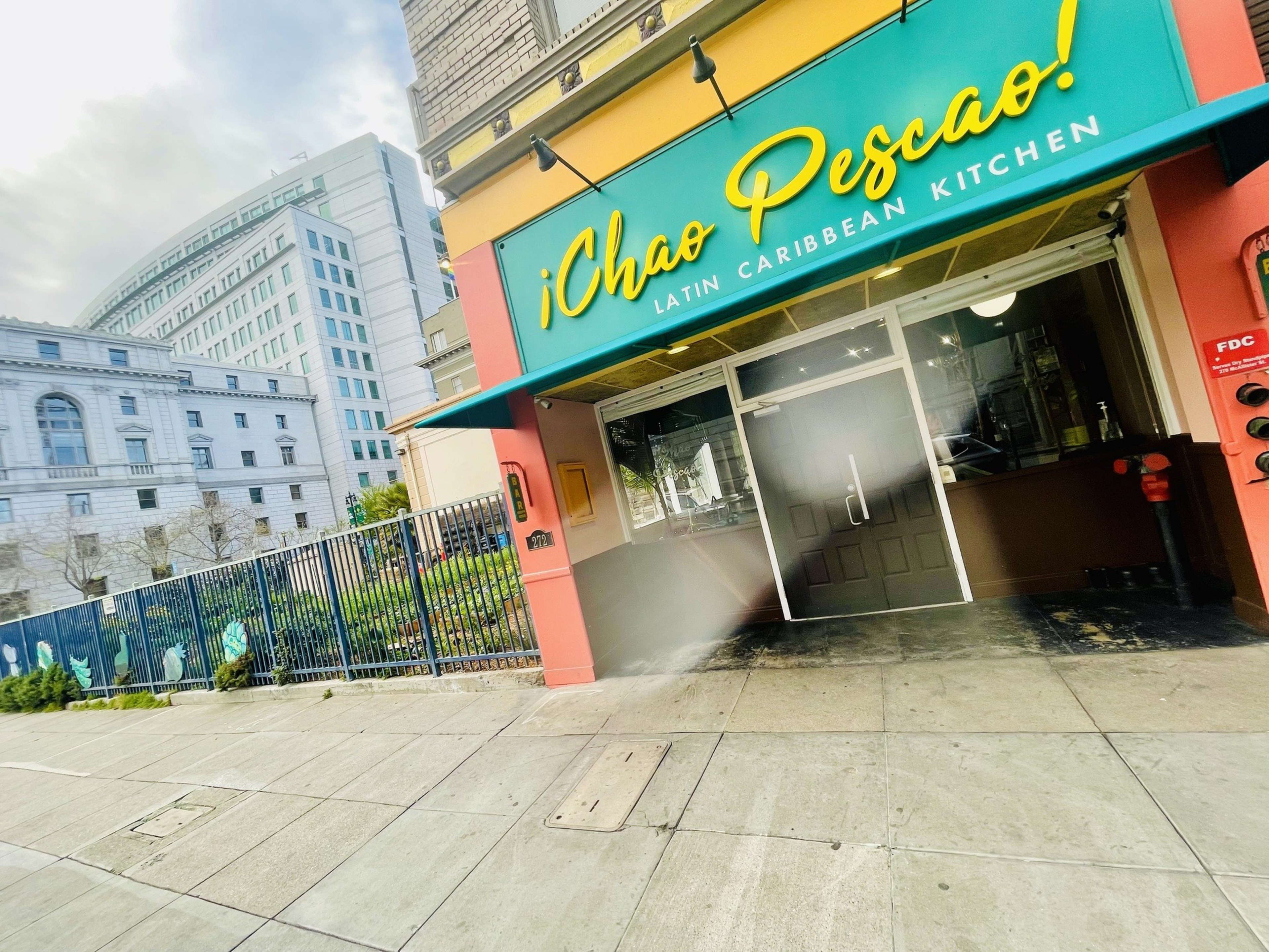 The image shows the entrance of &quot;¡Chao Pescao! Latin Caribbean Kitchen&quot; with colorful signage, a building in the background, and a fenced sidewalk.