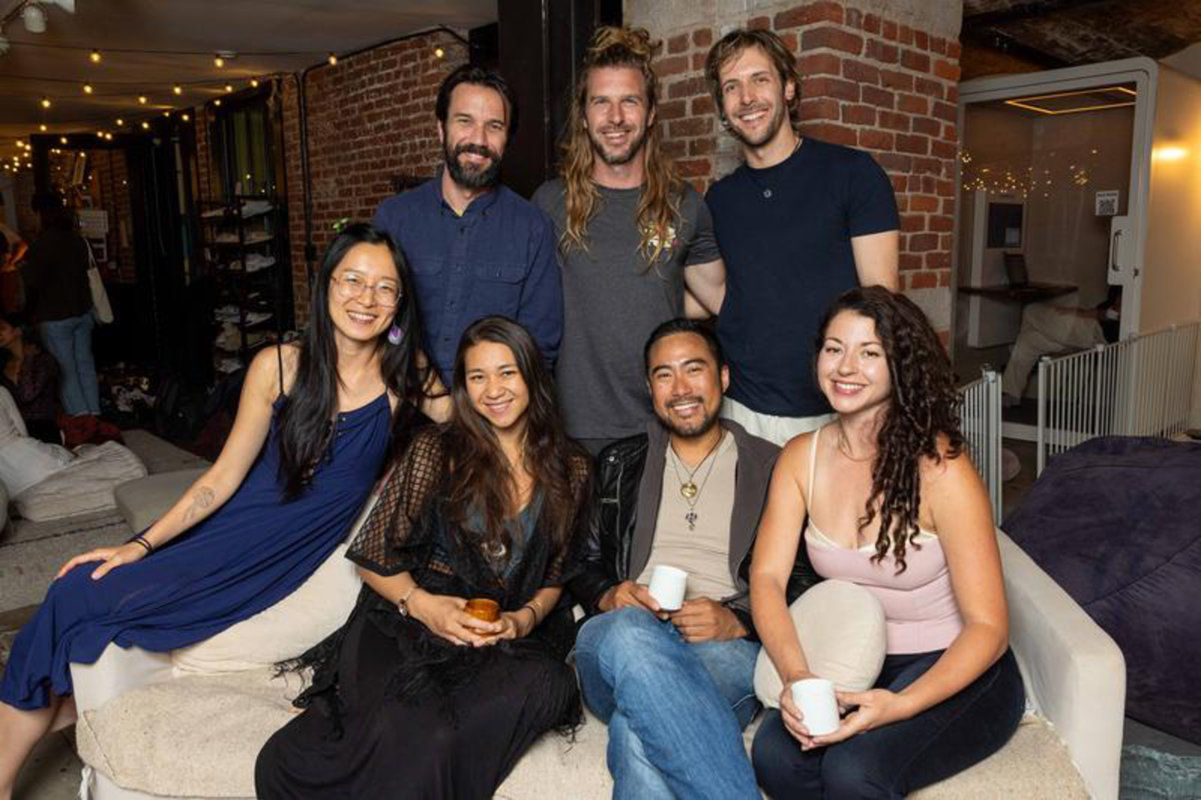 Seven people, three men and four women, smile while sitting and standing in a cozy room with brick walls and string lights overhead.
