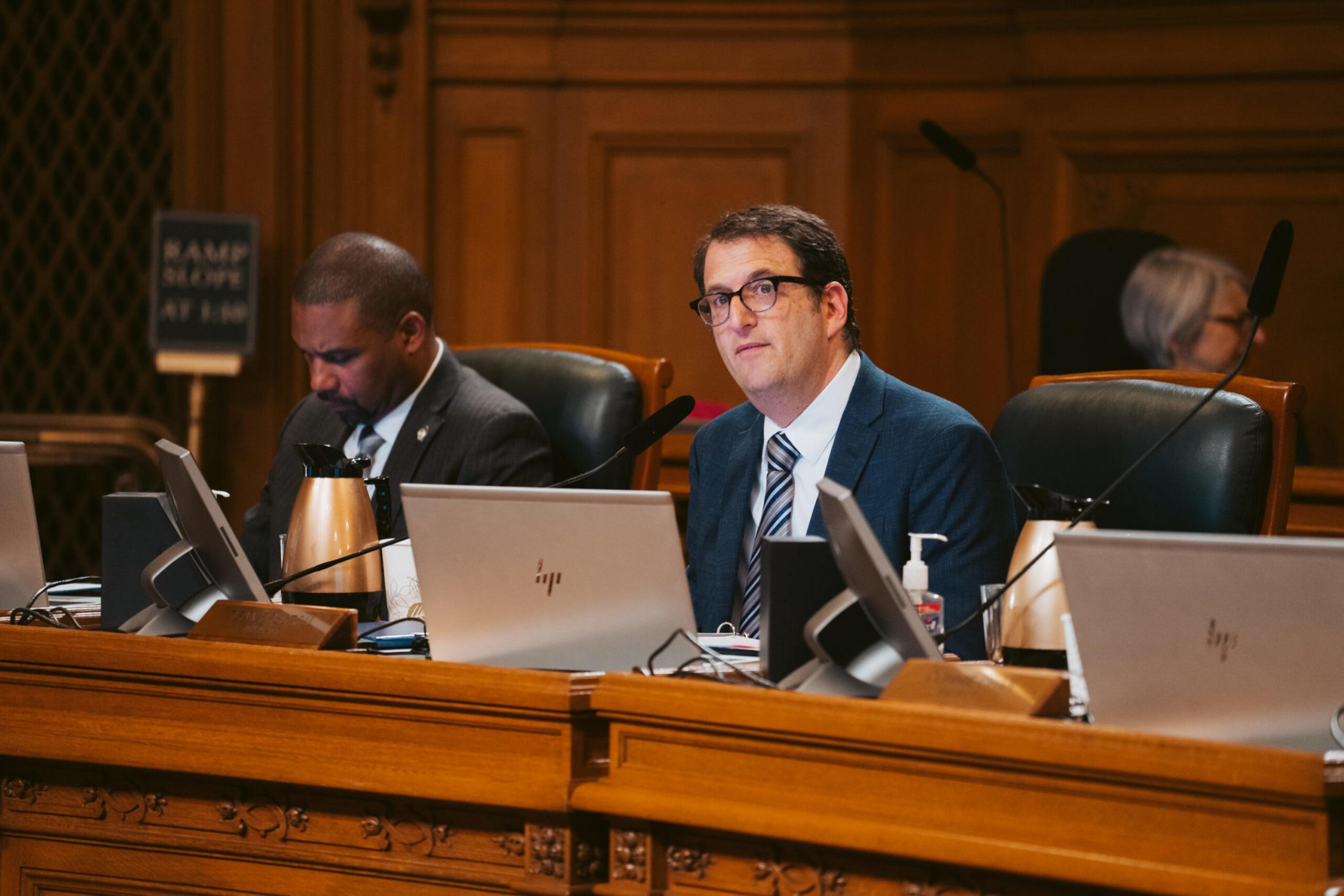 Two men in suits sit at a wooden conference table with laptops and microphones, with one man looking forward and the other looking down.