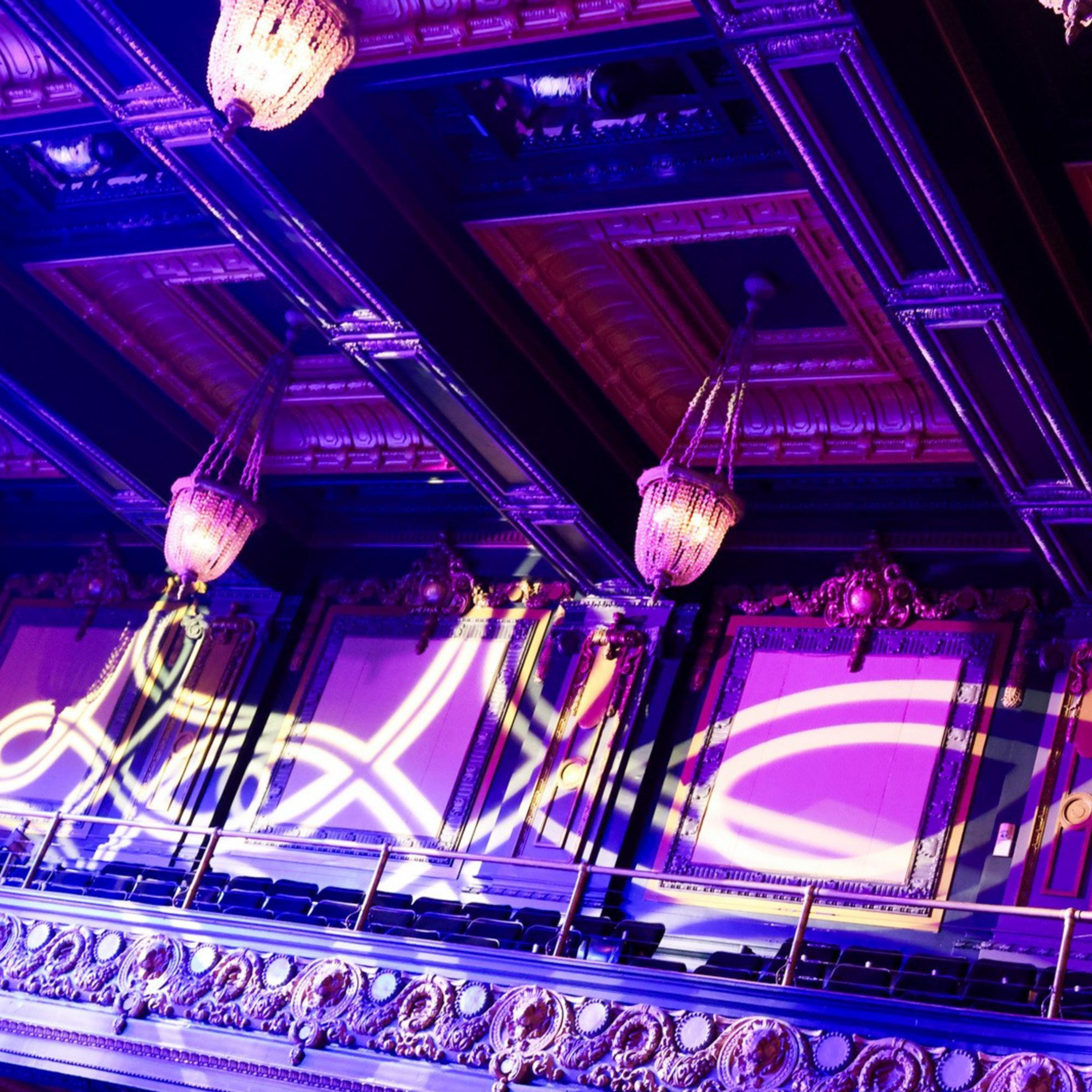The image shows an ornate balcony in a theater with intricate moldings, chandeliers, and purple lighting. Decorative light patterns are projected on the walls.