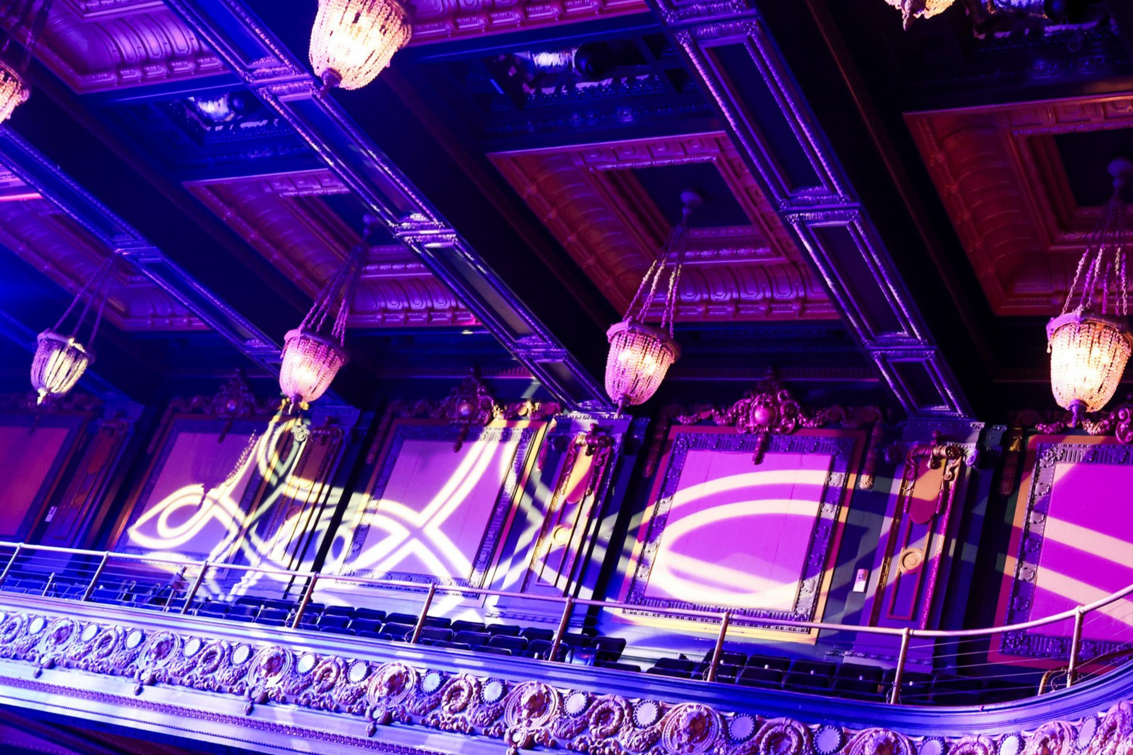 The image shows an ornate balcony in a theater with intricate moldings, chandeliers, and purple lighting. Decorative light patterns are projected on the walls.
