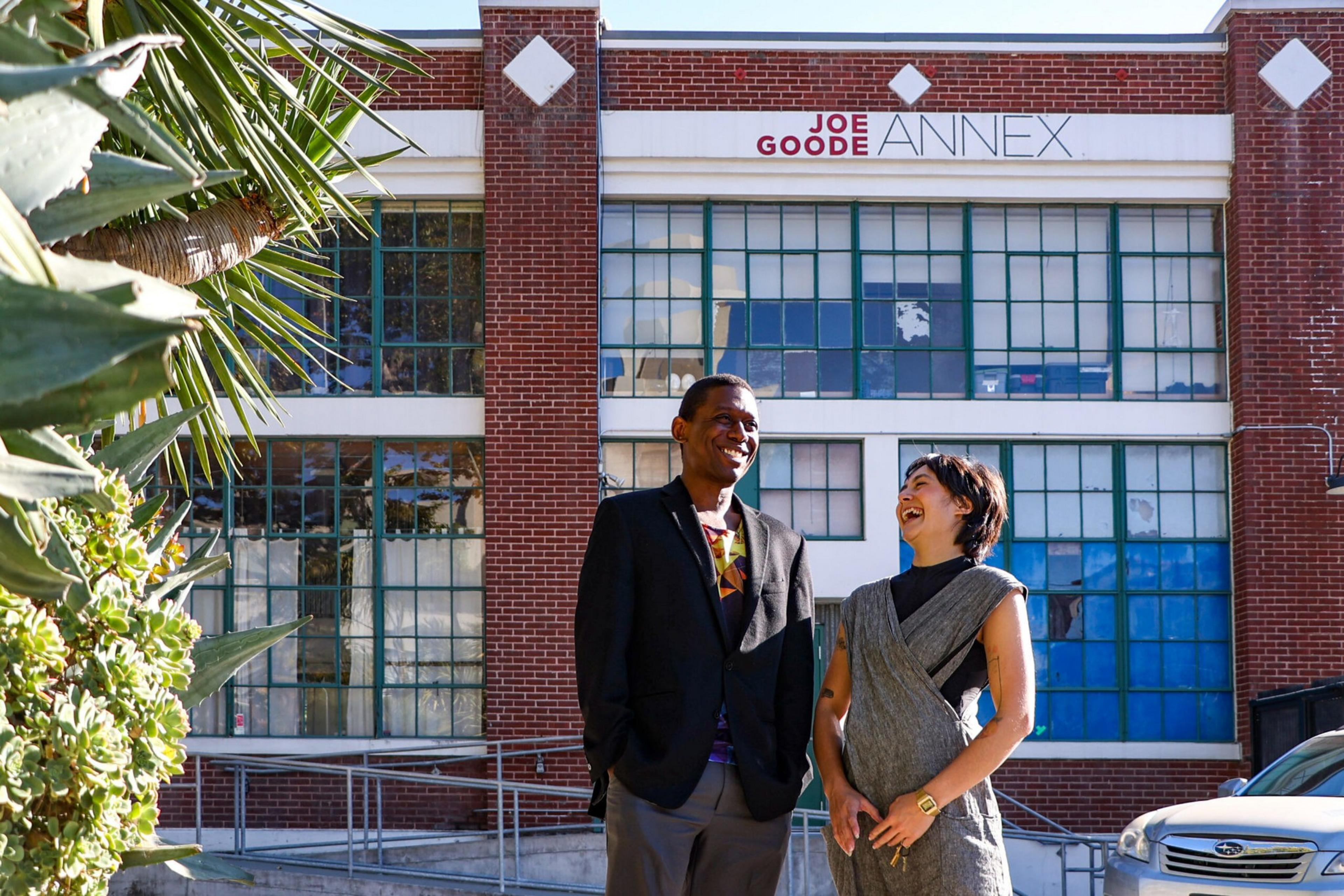 Two people stand laughing outside a brick building labeled "Joe Goode Annex," with large multi-paned windows and surrounding greenery.