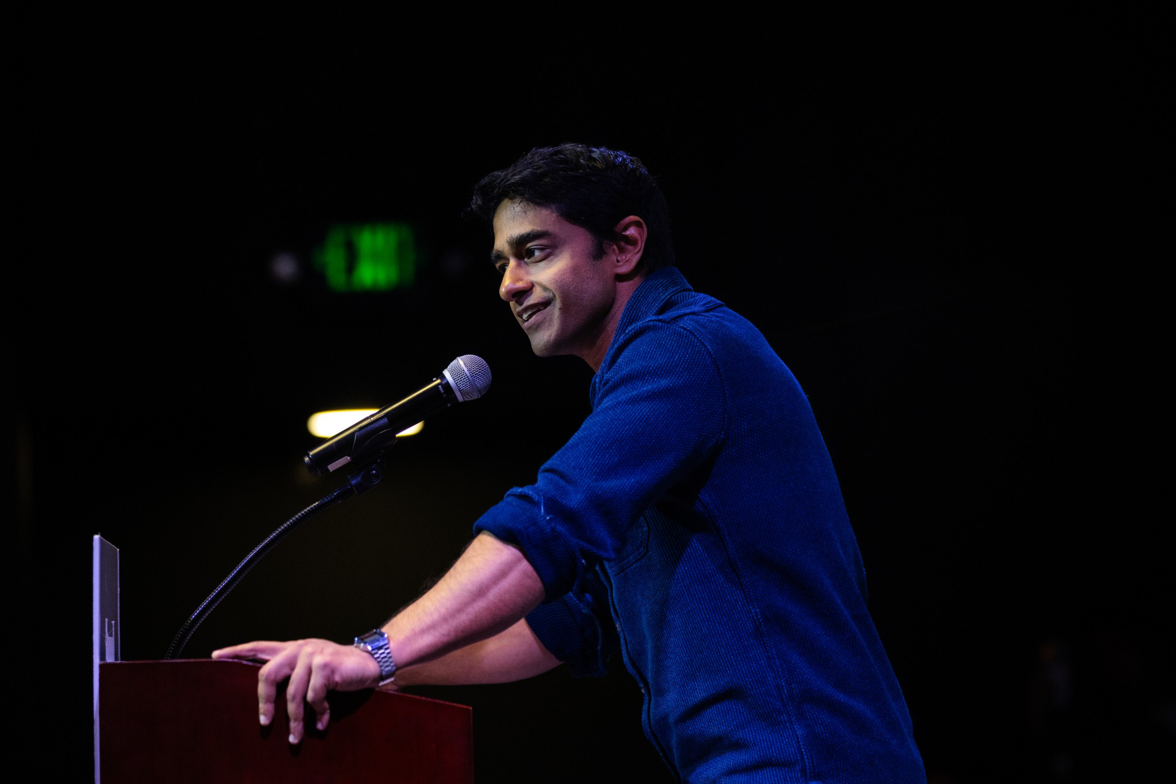 A man in a blue shirt speaks into a microphone while leaning on a podium, smiling against a dark background with a faint exit sign.
