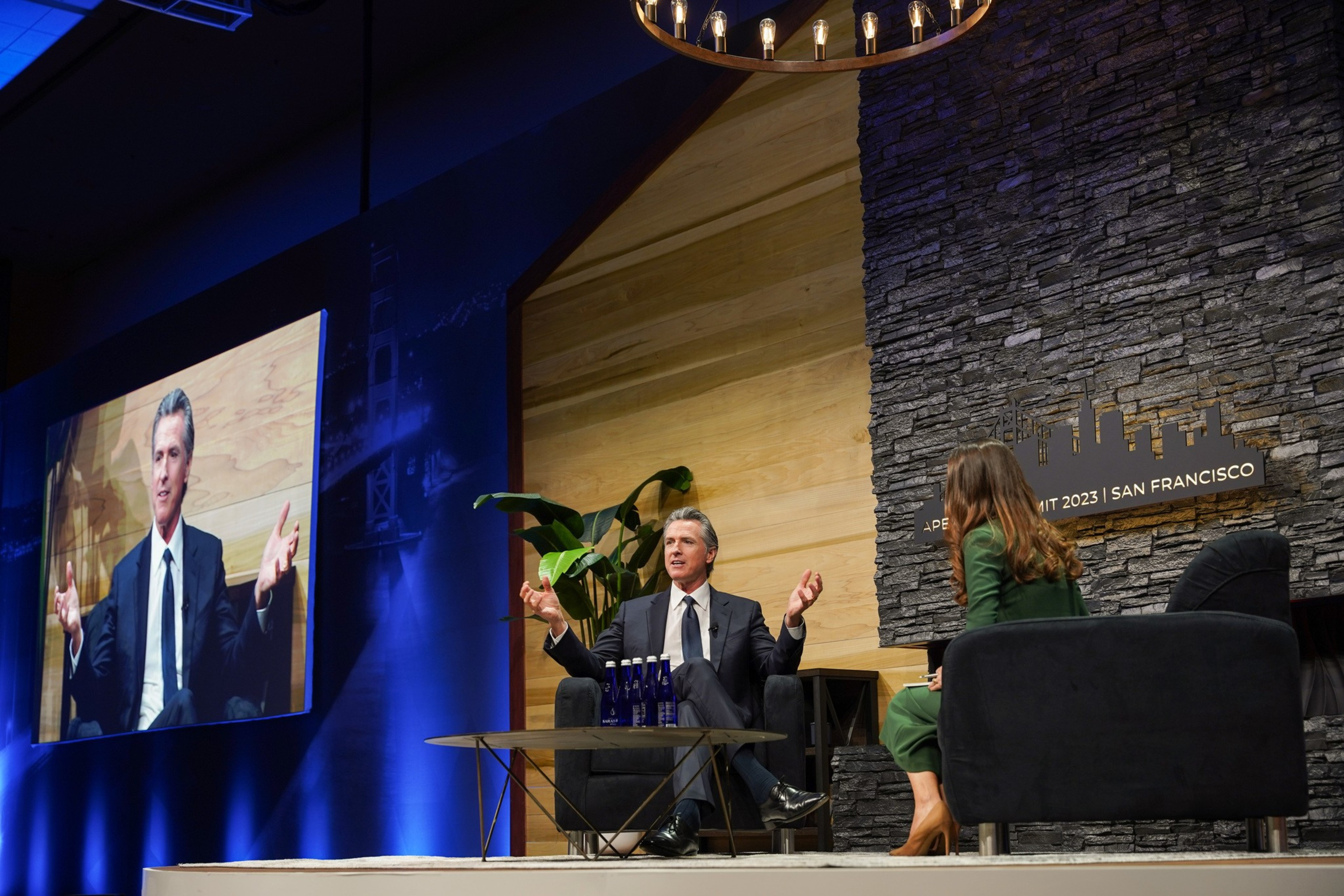 Gavin Newsom is seated in a chair with his arms outstretched and legs crossed. To his right, in a separate chair, is BBC presenter Helena Humphrey.