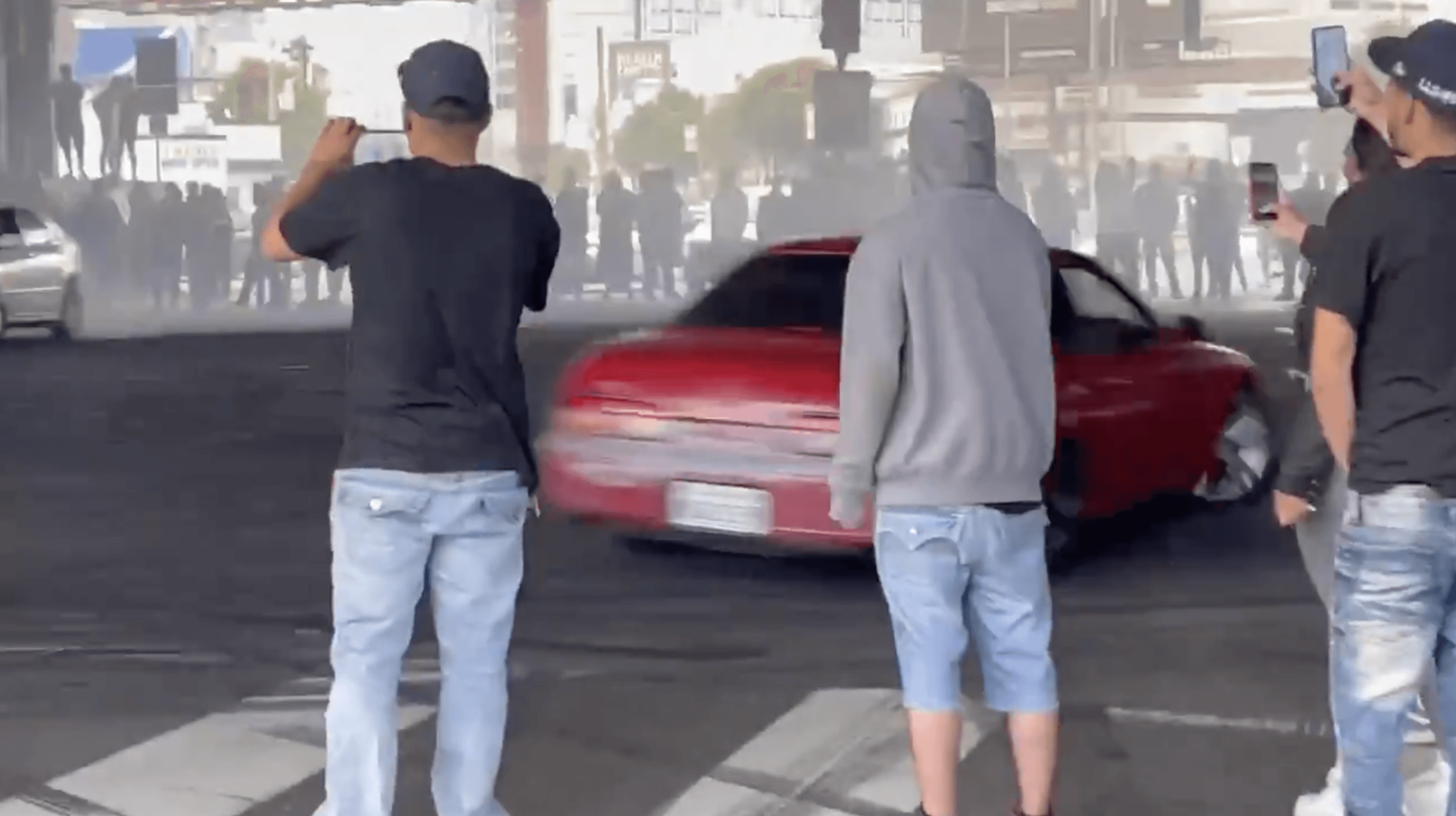 Three men stand watching and filming a red car drifting on a city street, with tire smoke and a crowd in the background.