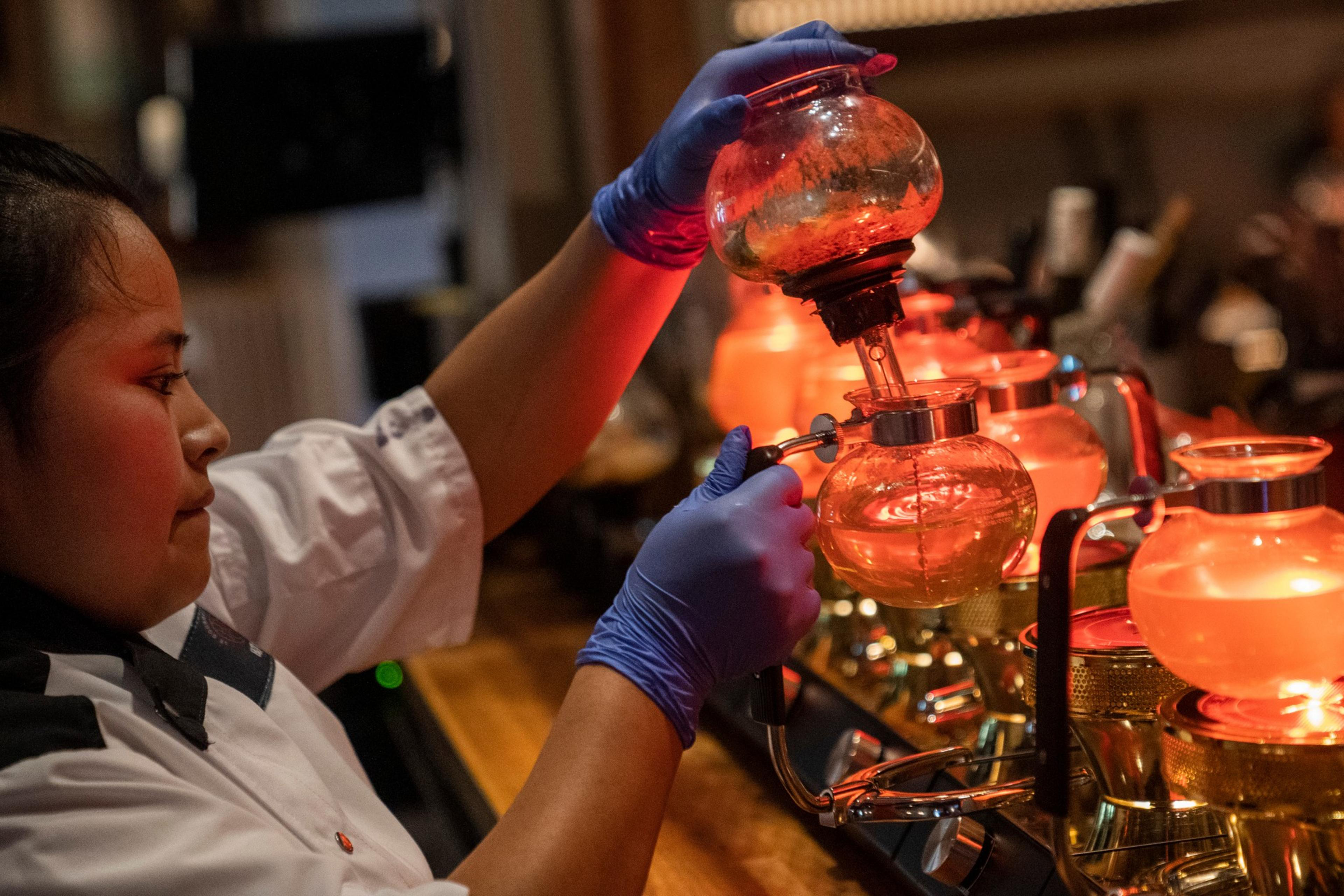 A person wearing blue gloves and a white coat uses a siphon coffee maker filled with illuminated glass globes, which glow in a warm orange hue.