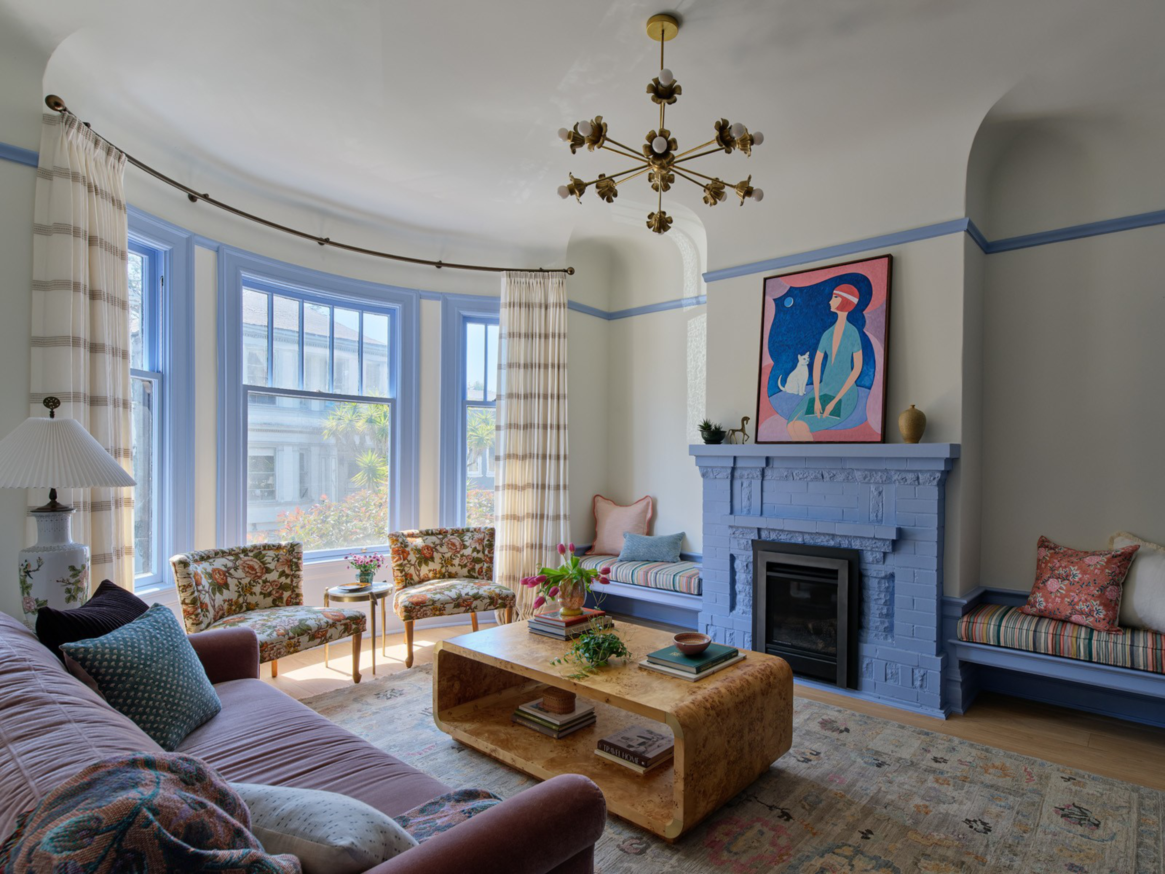 A cozy living room features a lavender couch, floral chairs, a marble coffee table, a blue-painted fireplace, built-in window seats, and colorful cushions.