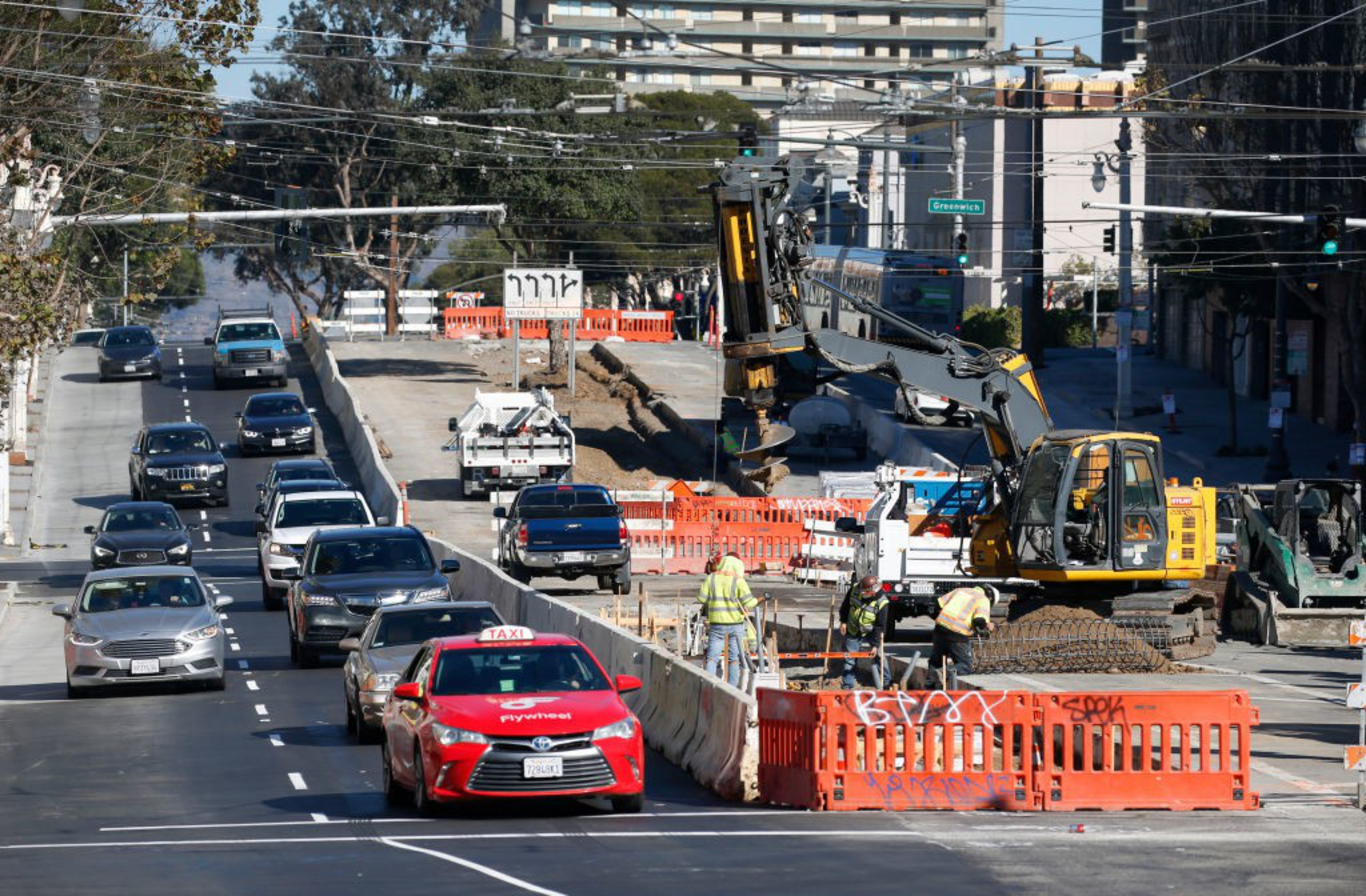 Vehicles drive on a multi-lane road next to a construction zone where workers use heavy machinery and orange barriers surround the site.