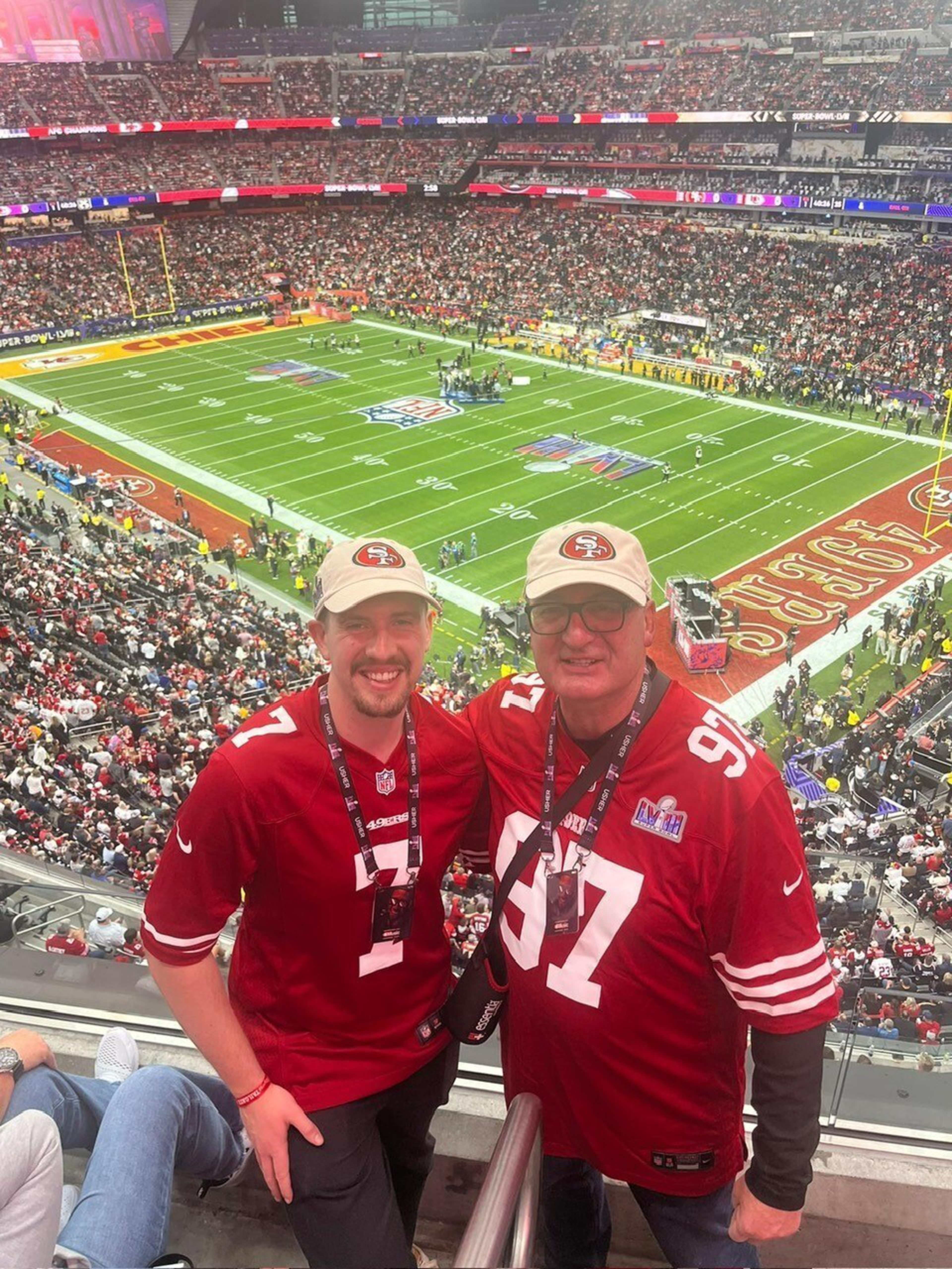 Two men wearing red San Francisco 49ers jerseys and caps stand smiling in a crowded stadium with an NFL football field below.