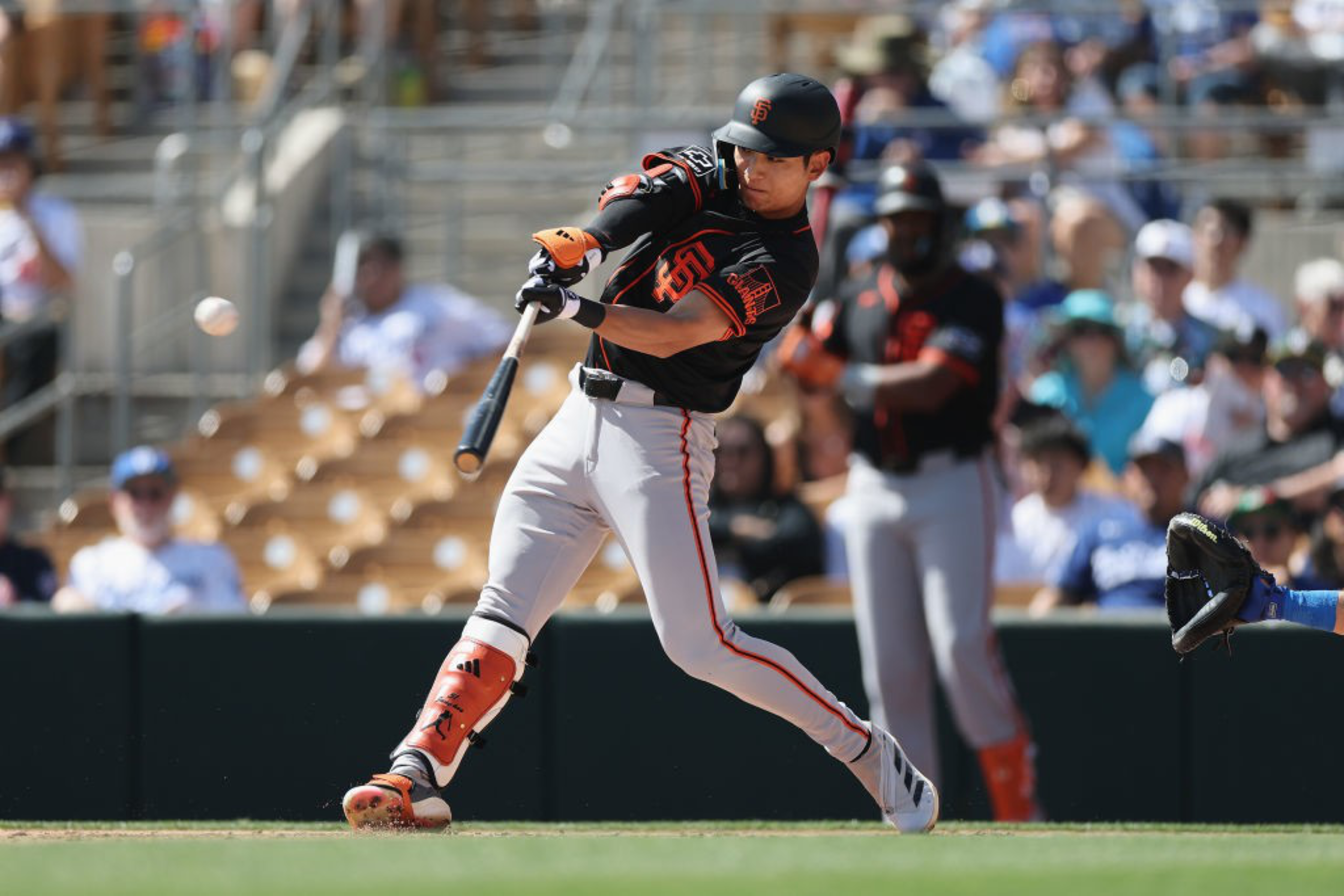 A baseball player in a black and orange uniform swings at a pitch, watched by a blurred crowd in stadium seats.