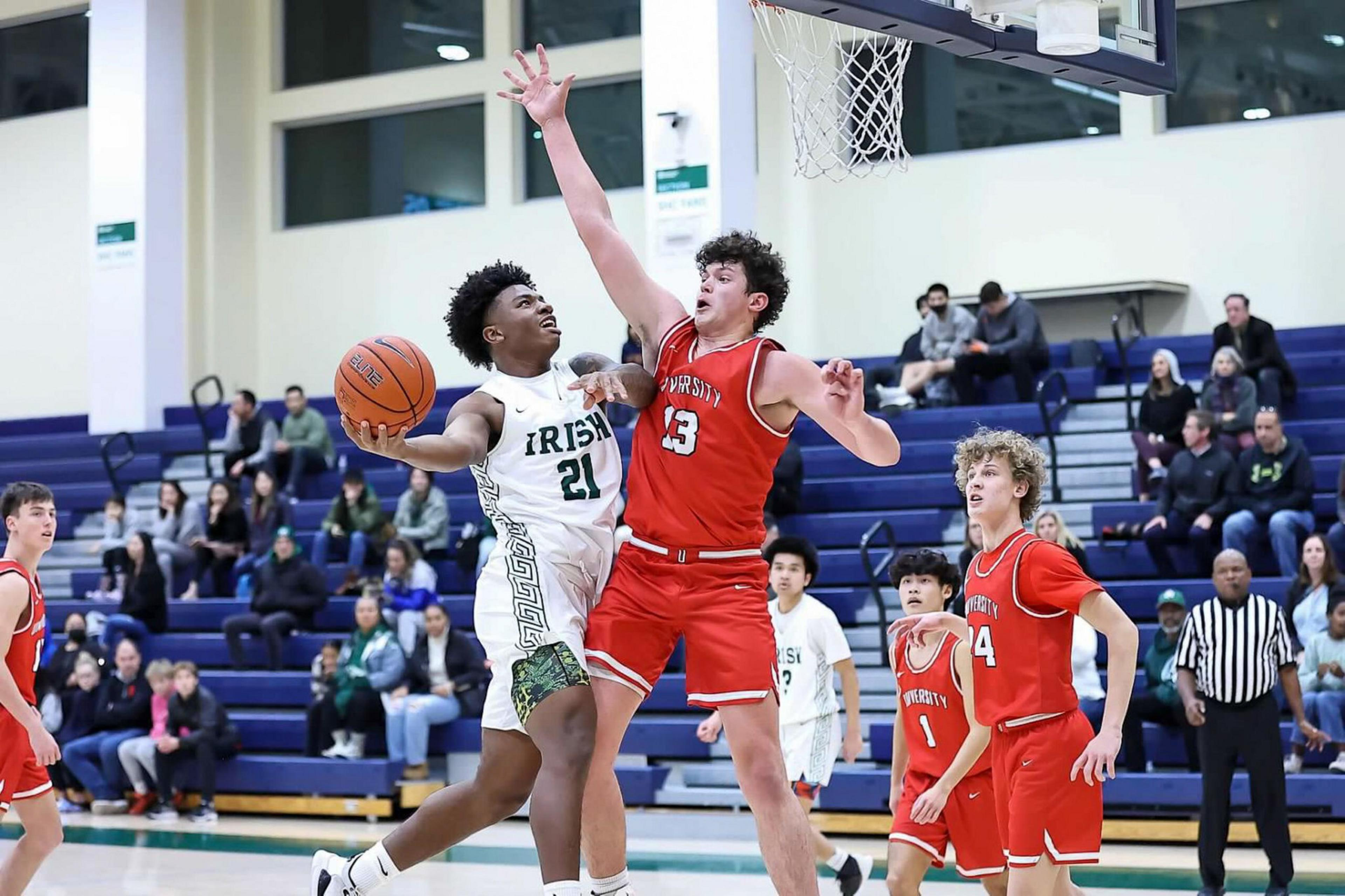 A basketball player in white with "Irish" on his jersey jumps to shoot while a red team defender with arms raised tries to block the shot near the basket.