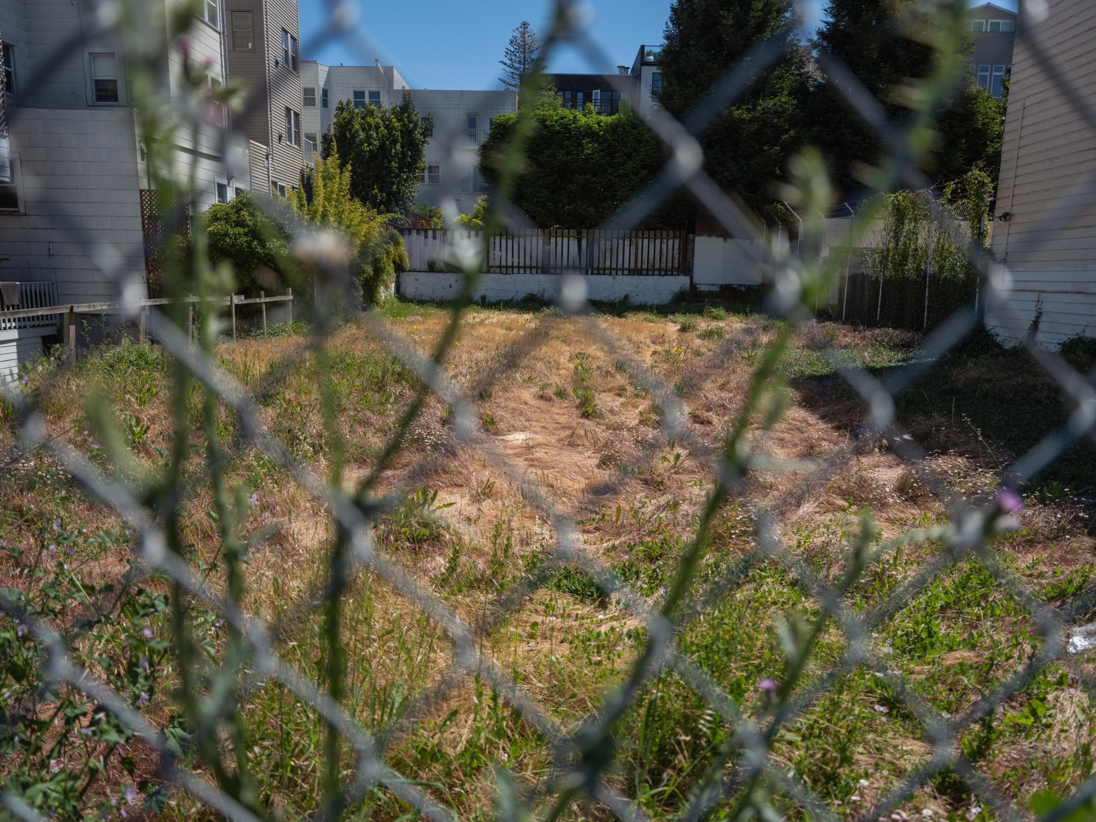 The image shows an overgrown vacant lot with dry grass, surrounded by a chain-link fence. Buildings and trees are visible in the background.
