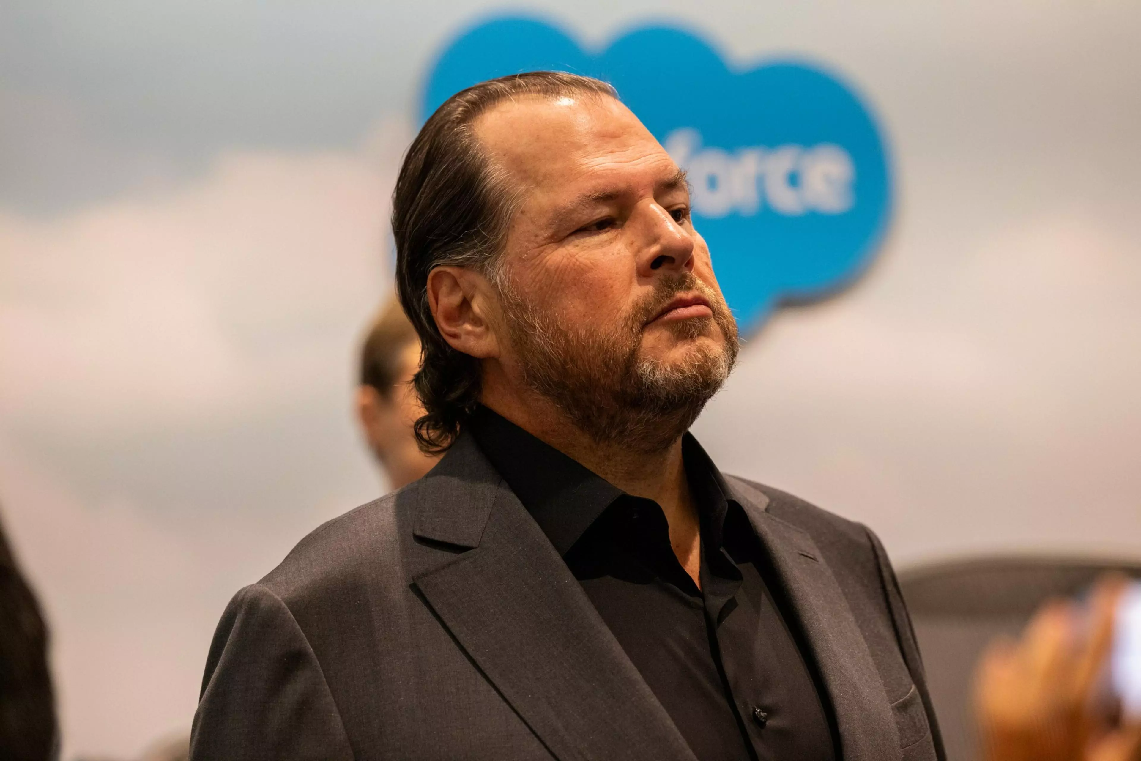 A man with slicked-back hair and a beard wears a dark suit and shirt, standing confidently with a blurred Salesforce logo in the background.