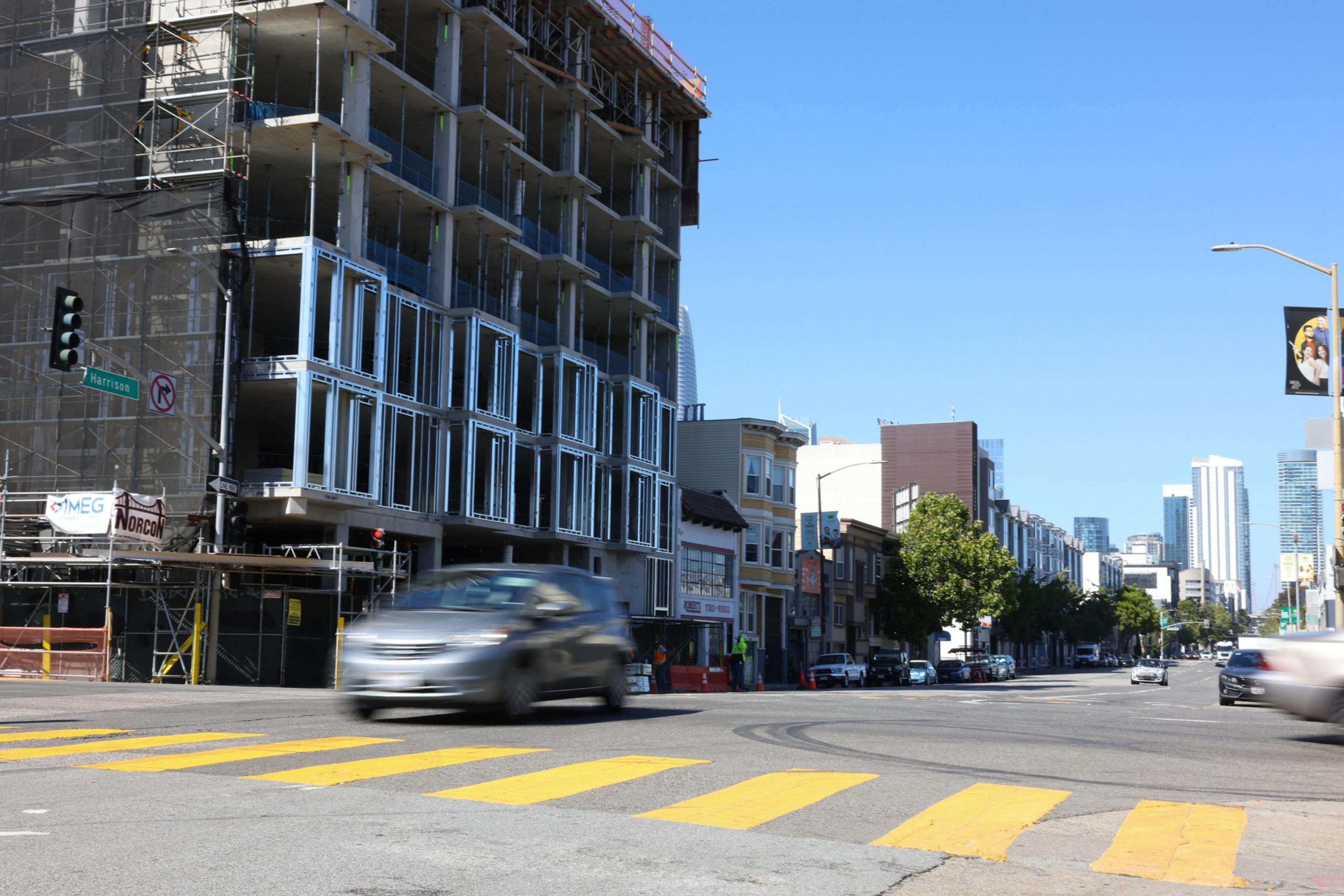 A busy urban street corner features a building under construction, moving cars, yellow crosswalk lines, and clear blue skies.