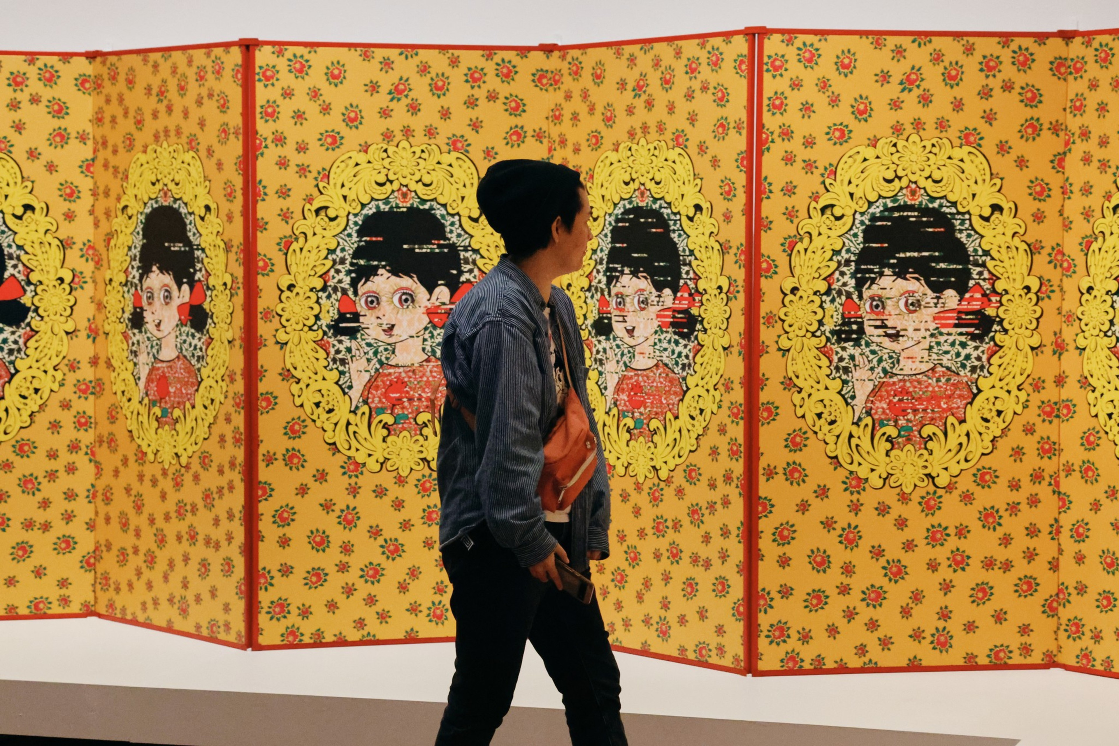 A person walks past an art installation featuring repeated cartoon portraits of a girl with black hair, red bows, and a red dress on a yellow floral background.